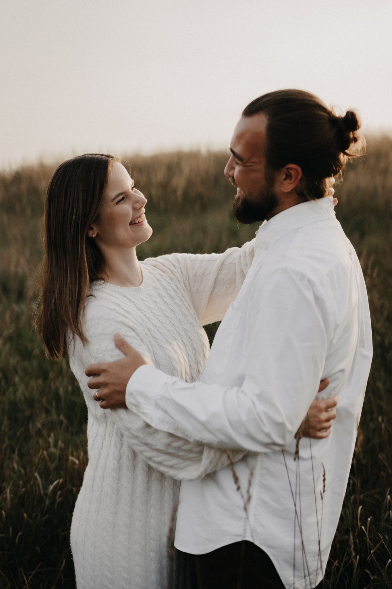Smiling couple embracing face to face in a golden field at sunset, both dressed in white.
