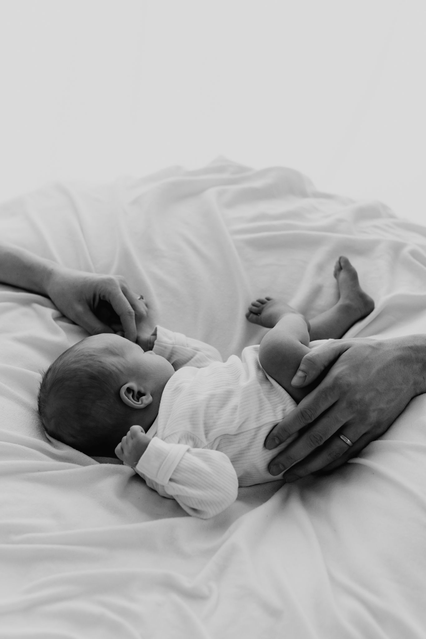Black and white image of parents hands tenderly holding a newborn baby on white sheets.