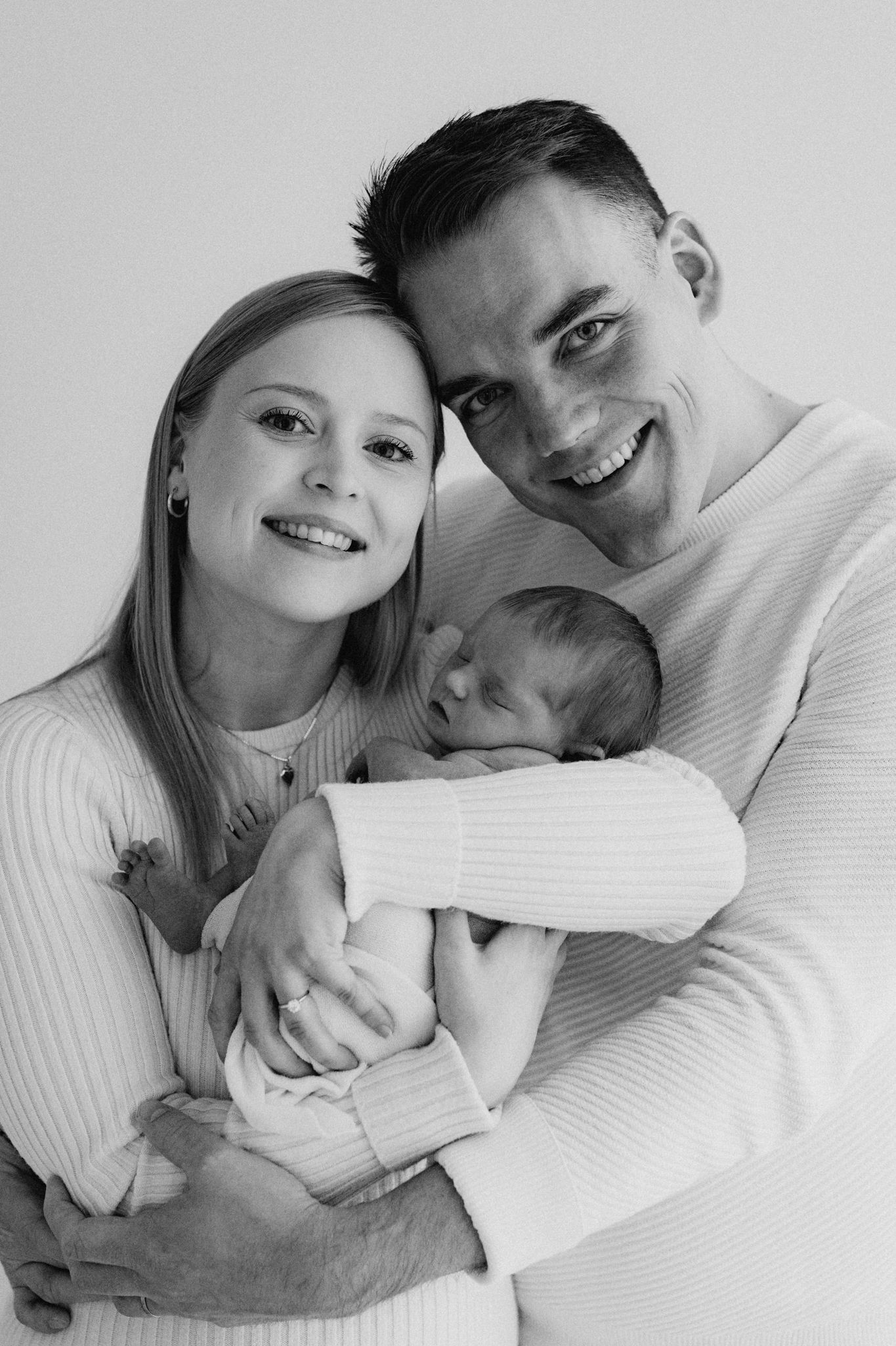 Black and white portrait of smiling parents holding their sleeping newborn baby.