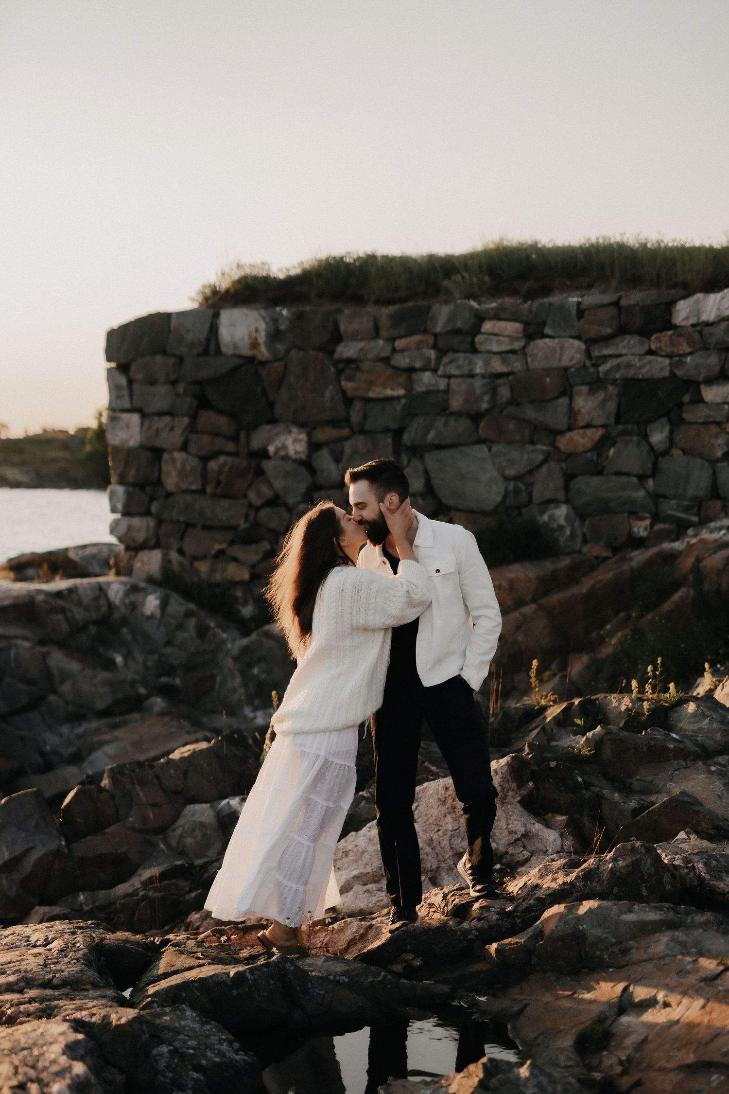 Couple sharing a tender kiss on rocky shore with ancient stone wall backdrop at sunset.