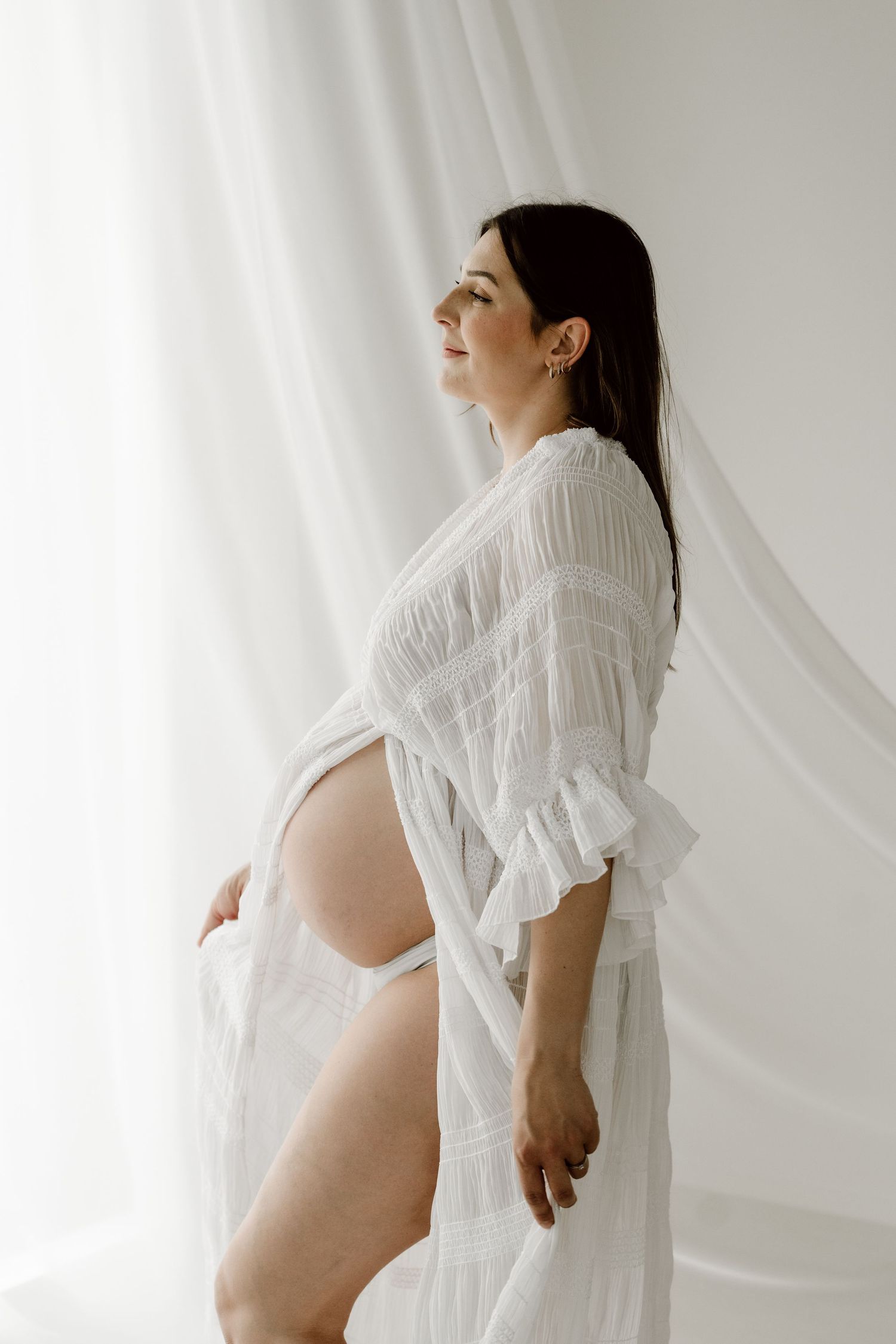 Raskaana oleva brunette nainen läpikuultavassa valkoisessa mekossa seisoo valkoisten verhojen vieressä pidellen vauvavatsaansa.