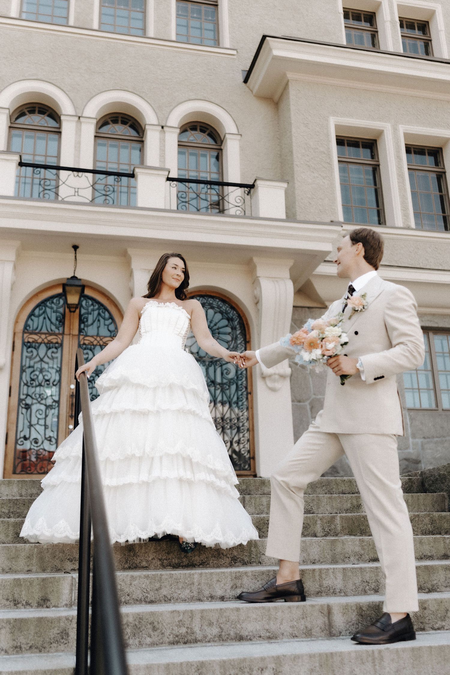 Wedding couple holding hands on steps of a classical building, groom holding a floral bouquet.