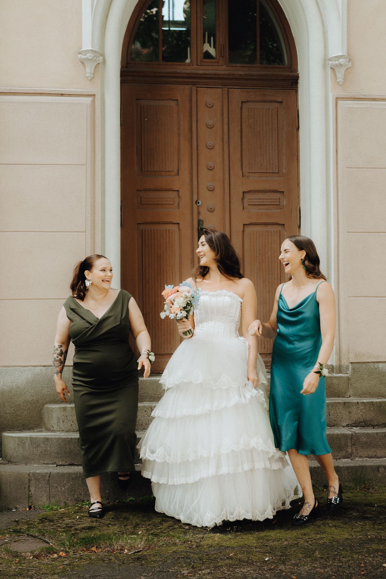 Bride in white tiered gown laughing with two bridesmaids in olive and teal dresses outside ornate wooden doors.