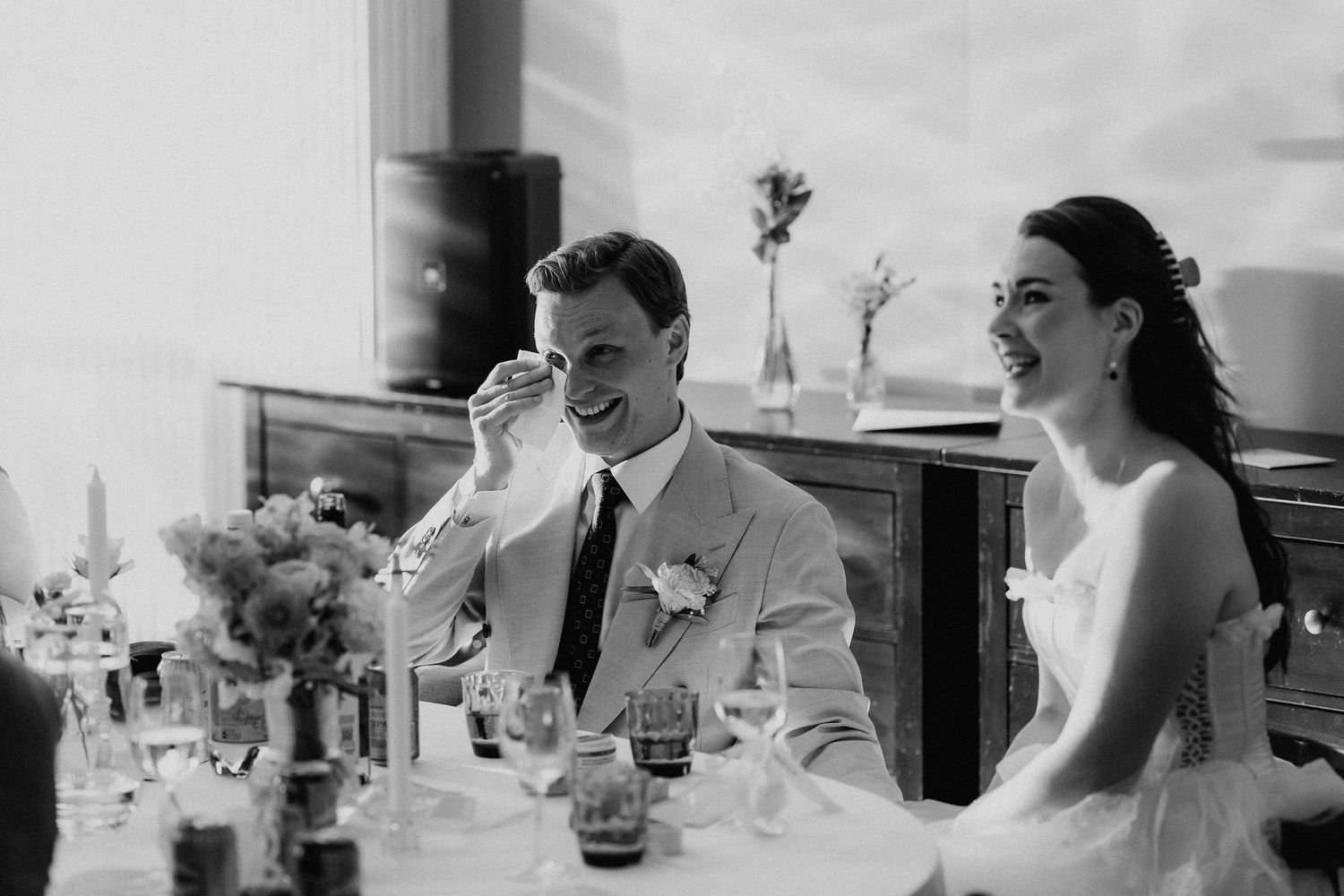 Black and white photo of bride and groom laughing joyfully during wedding reception speeches.