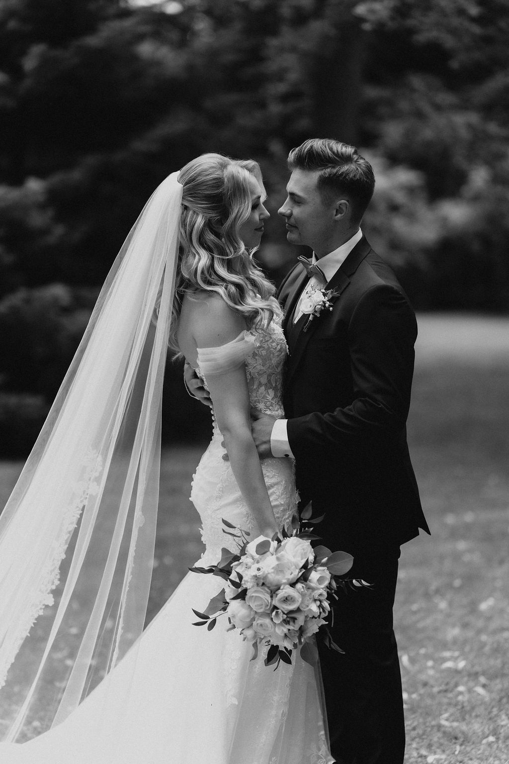 Black and white photo of bride and groom nearly kissing, bride holding white rose bouquet with greenery.