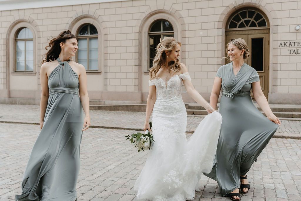 Bride in lace gown walks with two bridesmaids in flowing sage green dresses on cobblestone street.