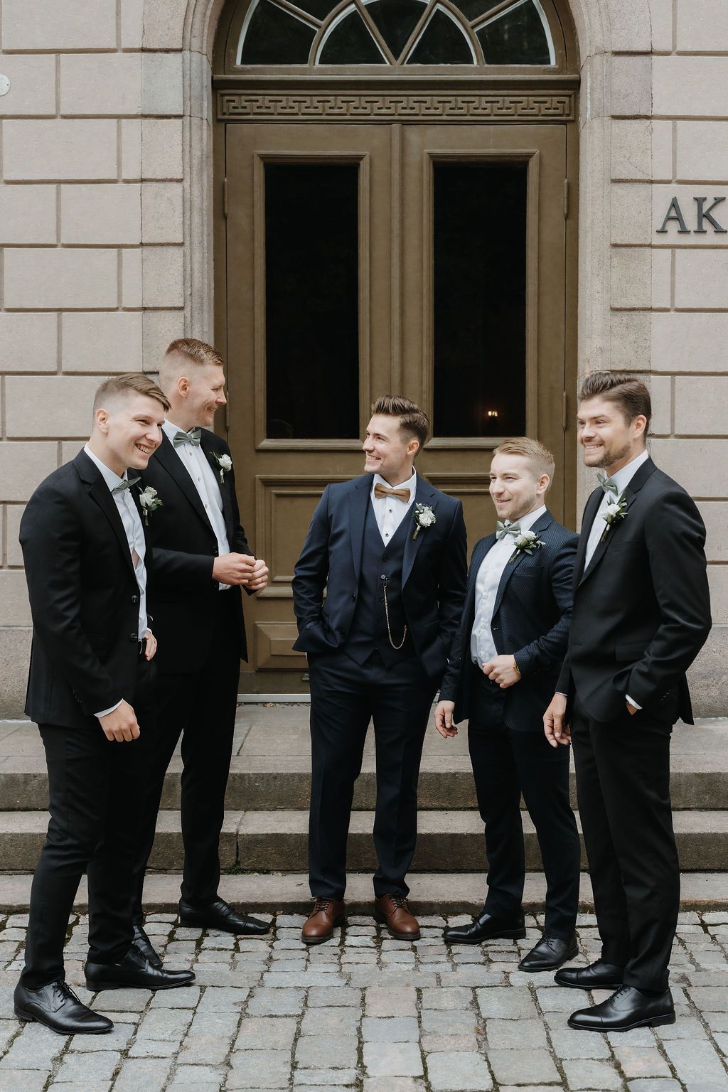 Five groomsmen in black and navy suits pose and smile outside a stone building.