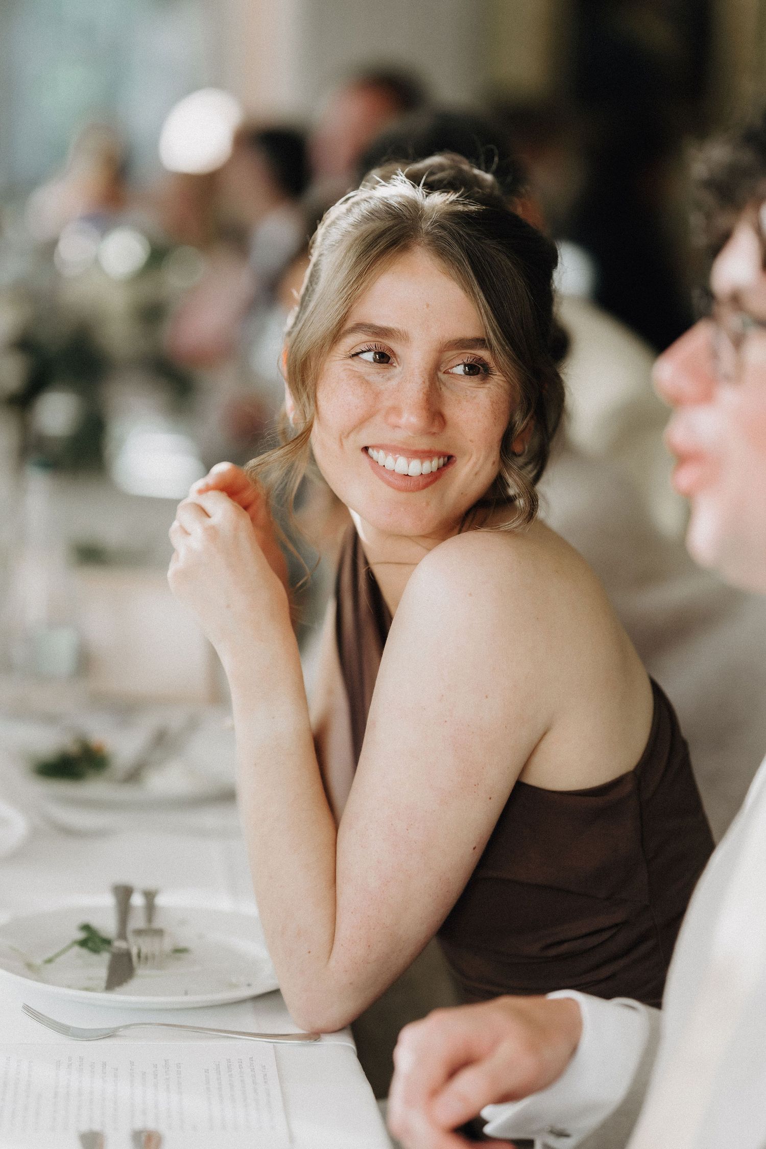 Smiling young woman in a brown dress enjoying a wedding reception dinner at an elegantly set table.