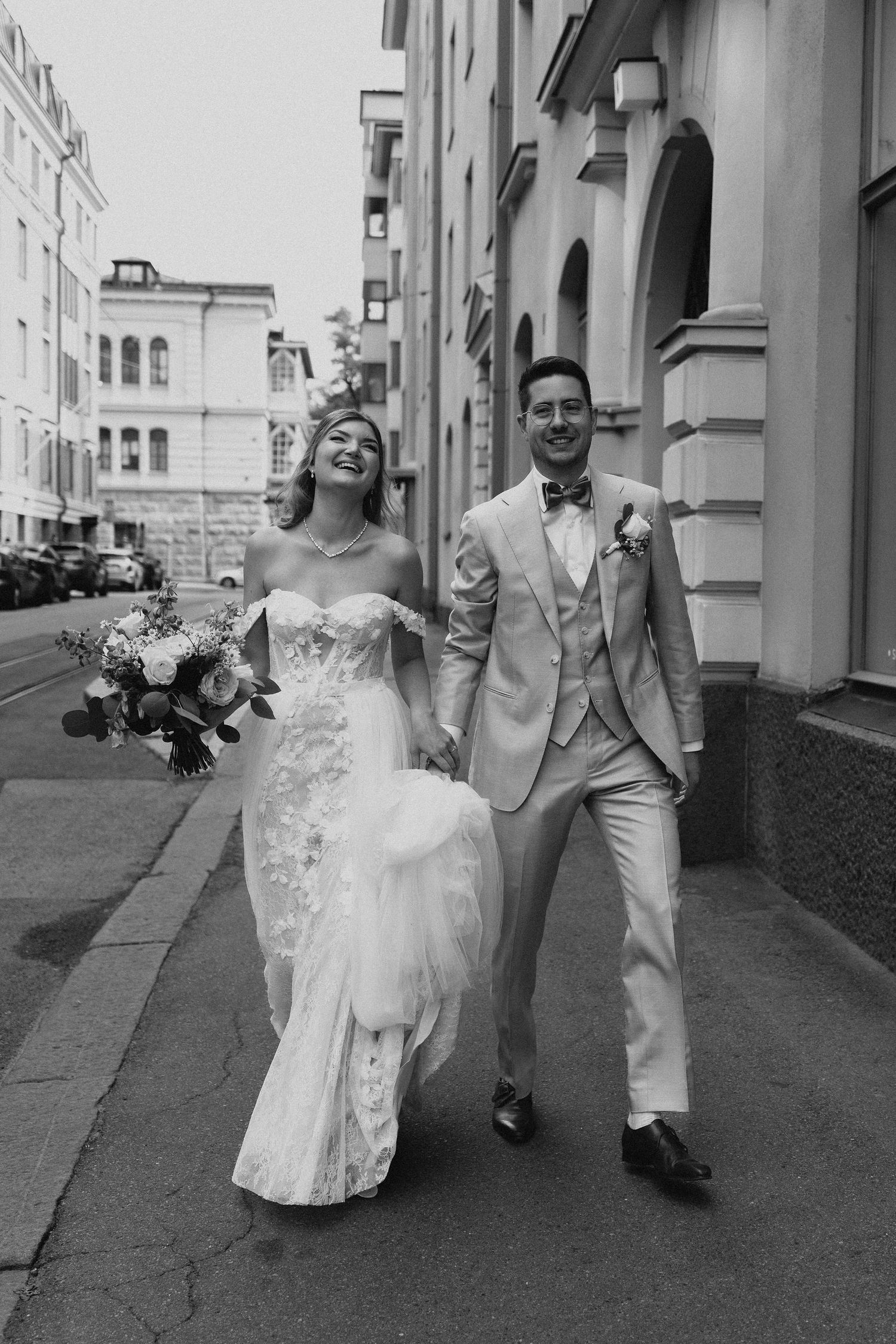 Black and white photo of smiling bride and groom walking hand in hand on a city street.