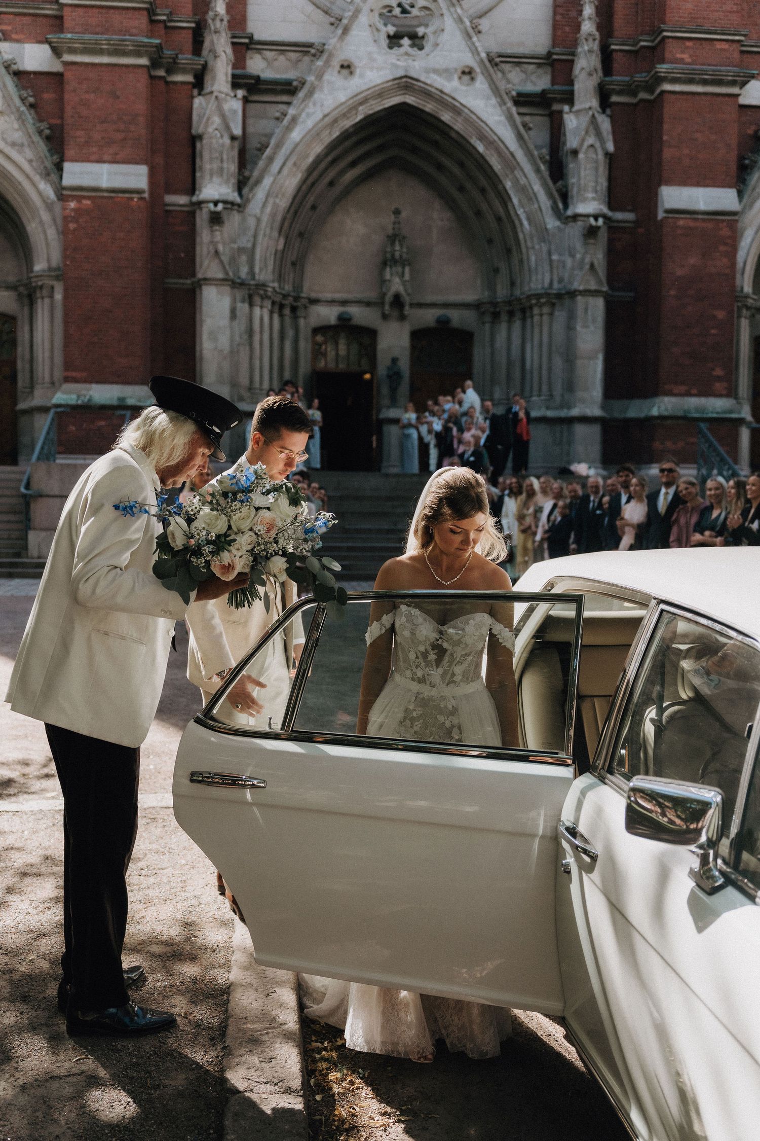 Bride in lace gown entering a vintage white Rolls-Royce outside a Gothic church with groom.