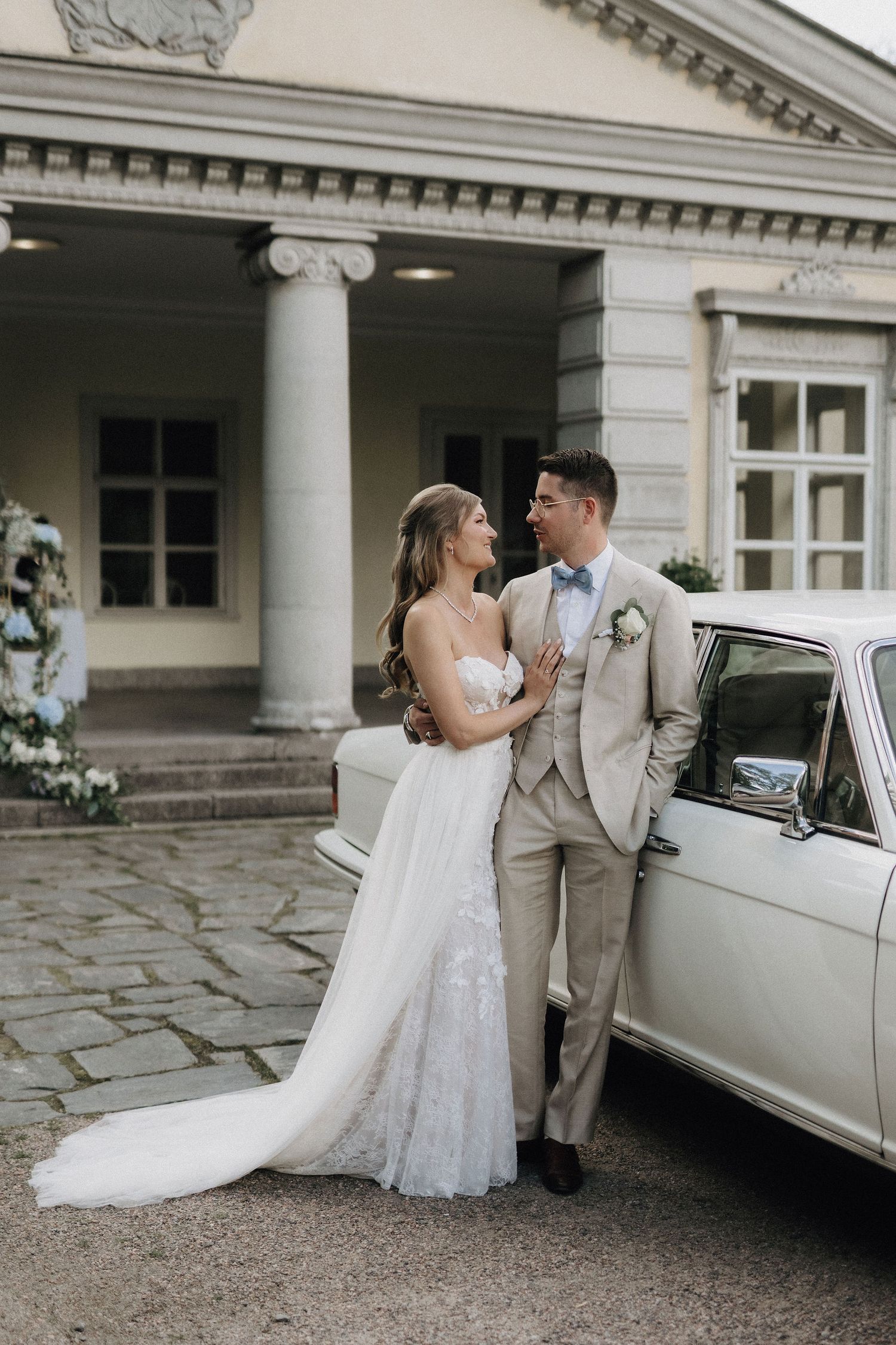 Bride in strapless lace gown and groom in beige suit posing beside a vintage white car at a manor.