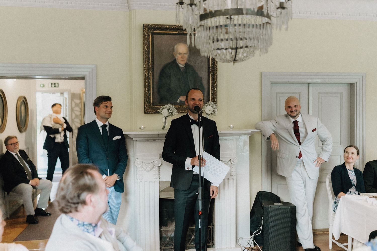 Wedding MC speaking at microphone in ornate room with chandelier and guests at decorated tables.