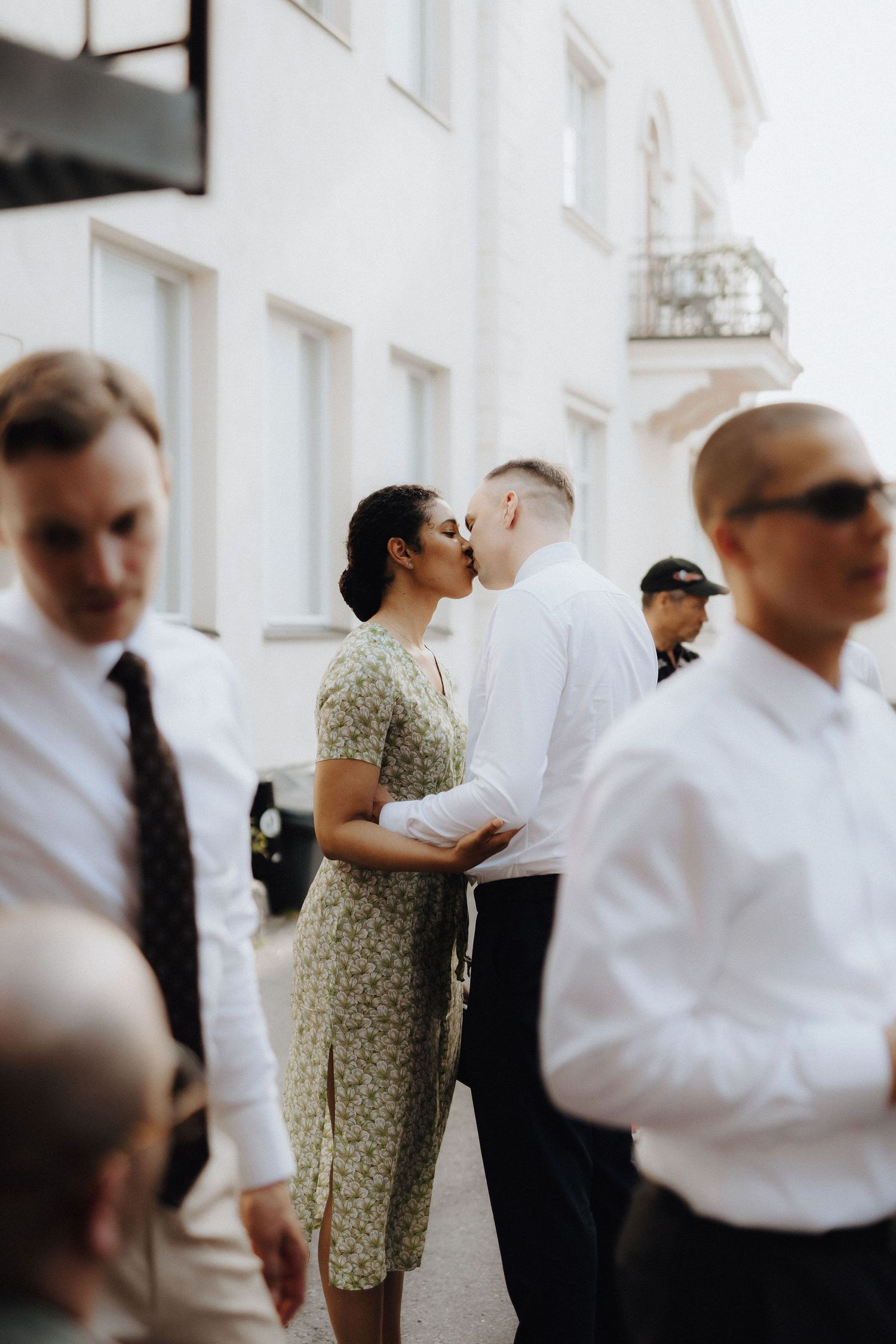 A couple shares a kiss outdoors surrounded by wedding guests in formal attire.