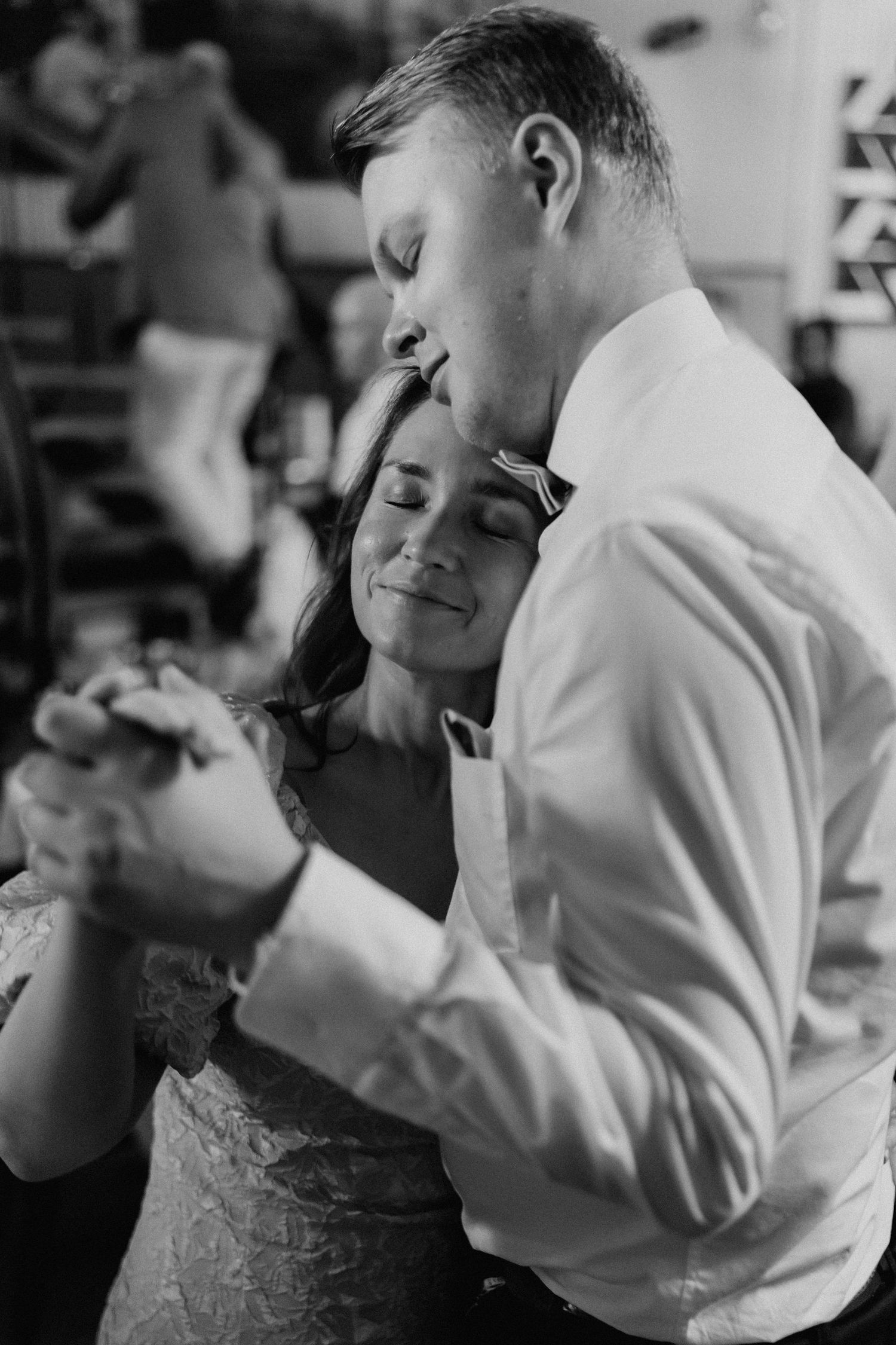 Black and white photo of groom kissing bride on forehead tenderly during slow dance at reception.