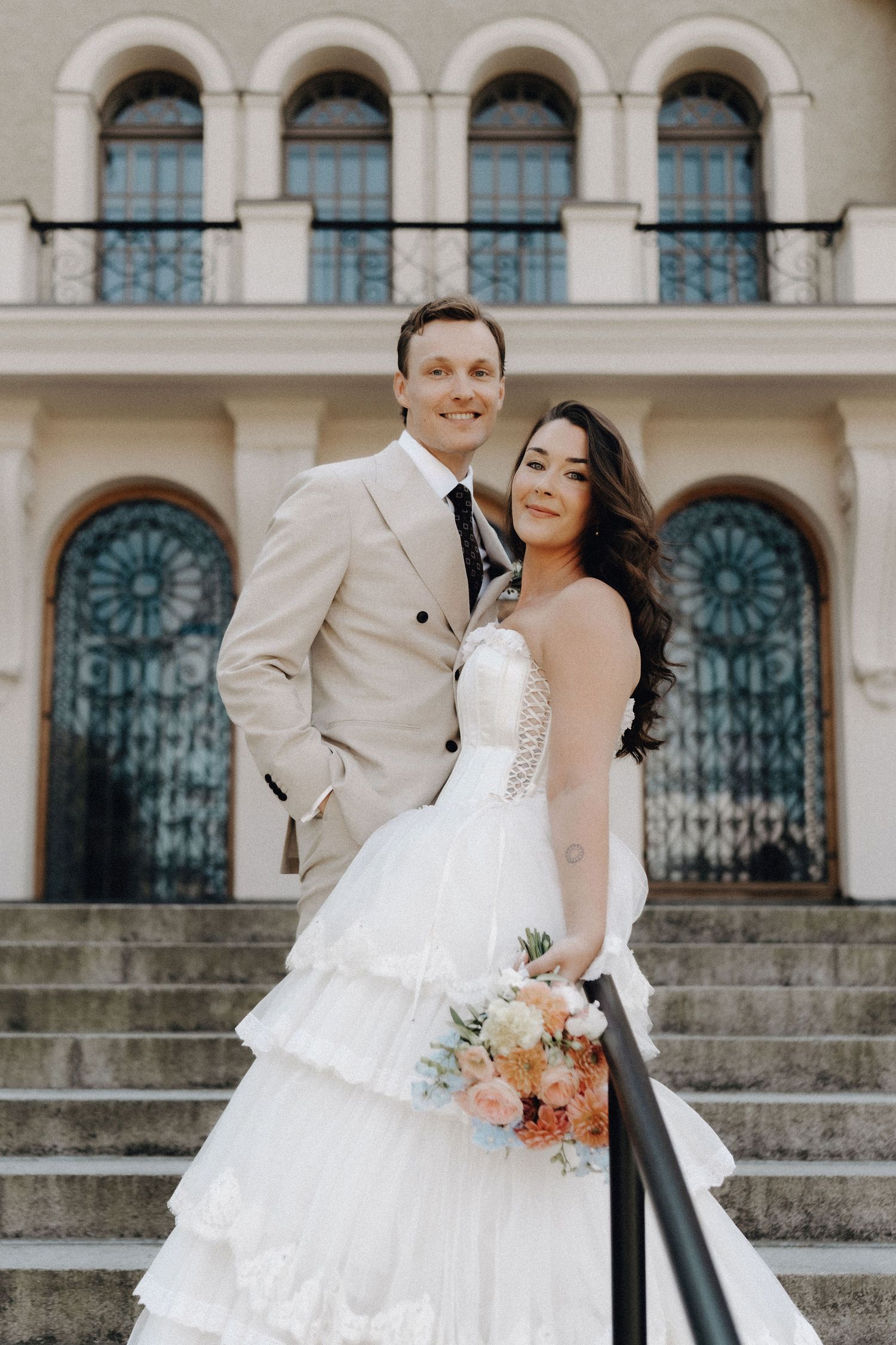 Smiling bride and groom posing together on steps before ornate arched windows of a grand building.