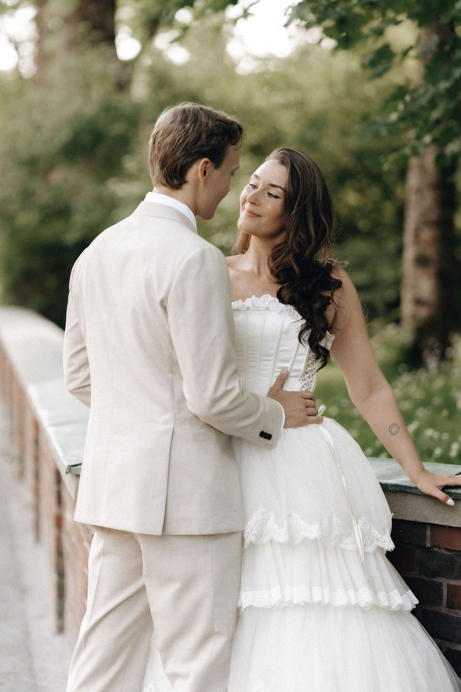 Bride in strapless tiered white gown and groom in ivory suit embracing in lush garden.