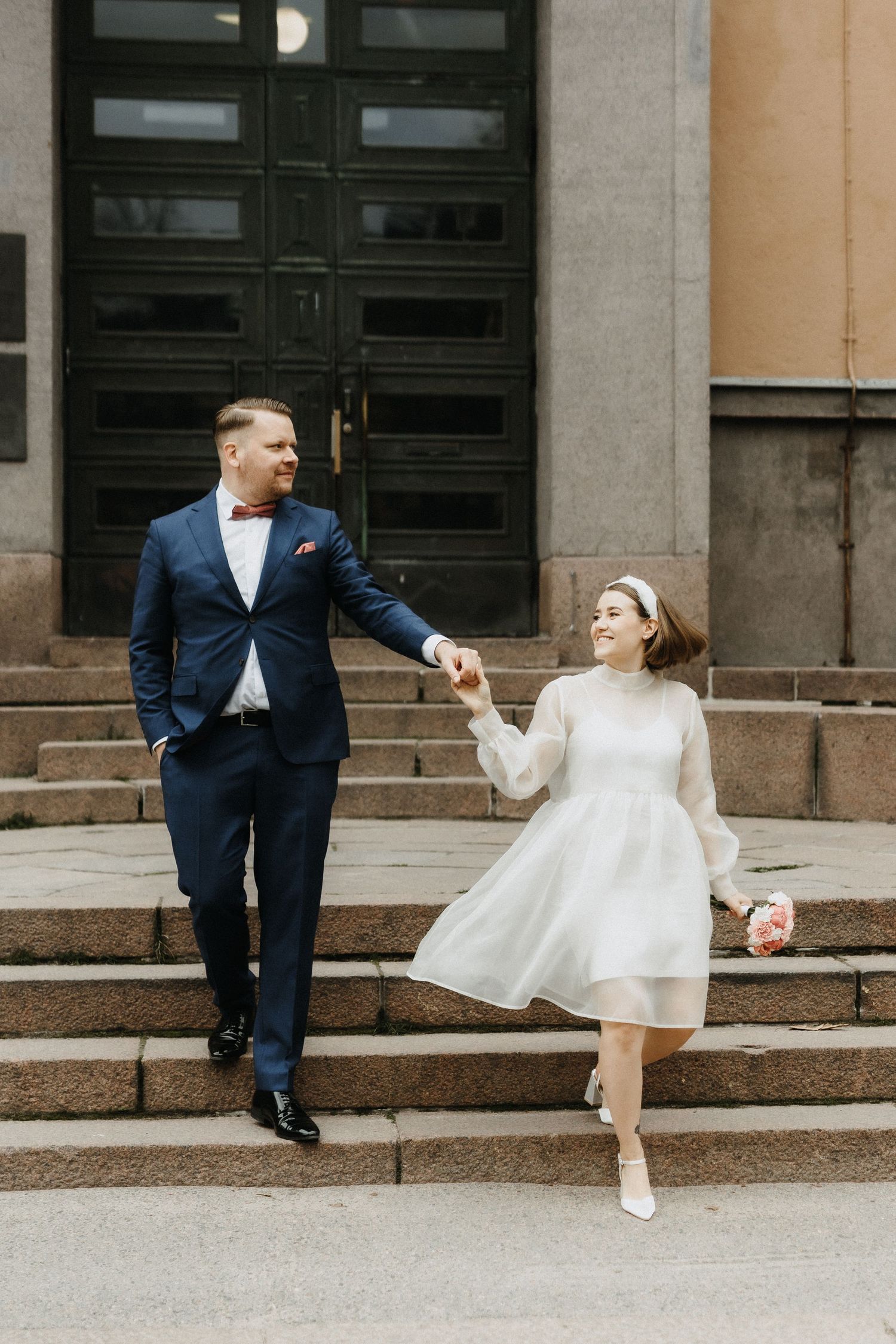Bride in short white dress and headband holding bouquet, joyfully walking down steps with groom in blue suit.