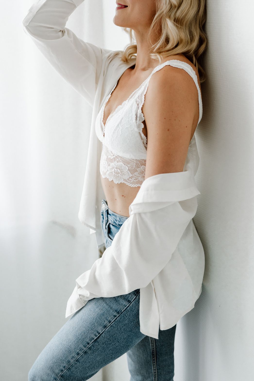 Blonde woman in white lace bralette and open shirt leaning against wall.