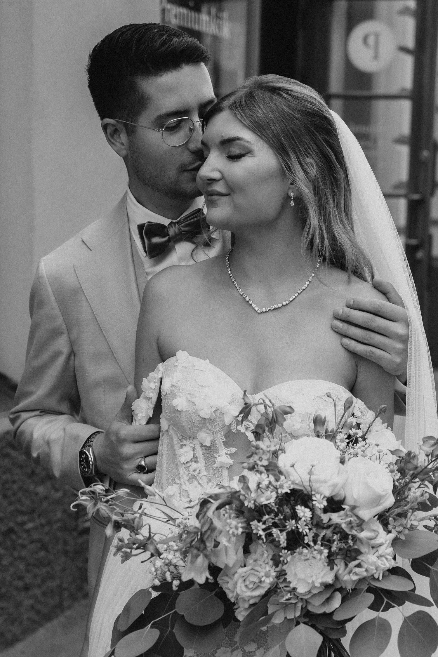 Black and white photo of groom kissing bride's temple, bride holding lush white floral bouquet.