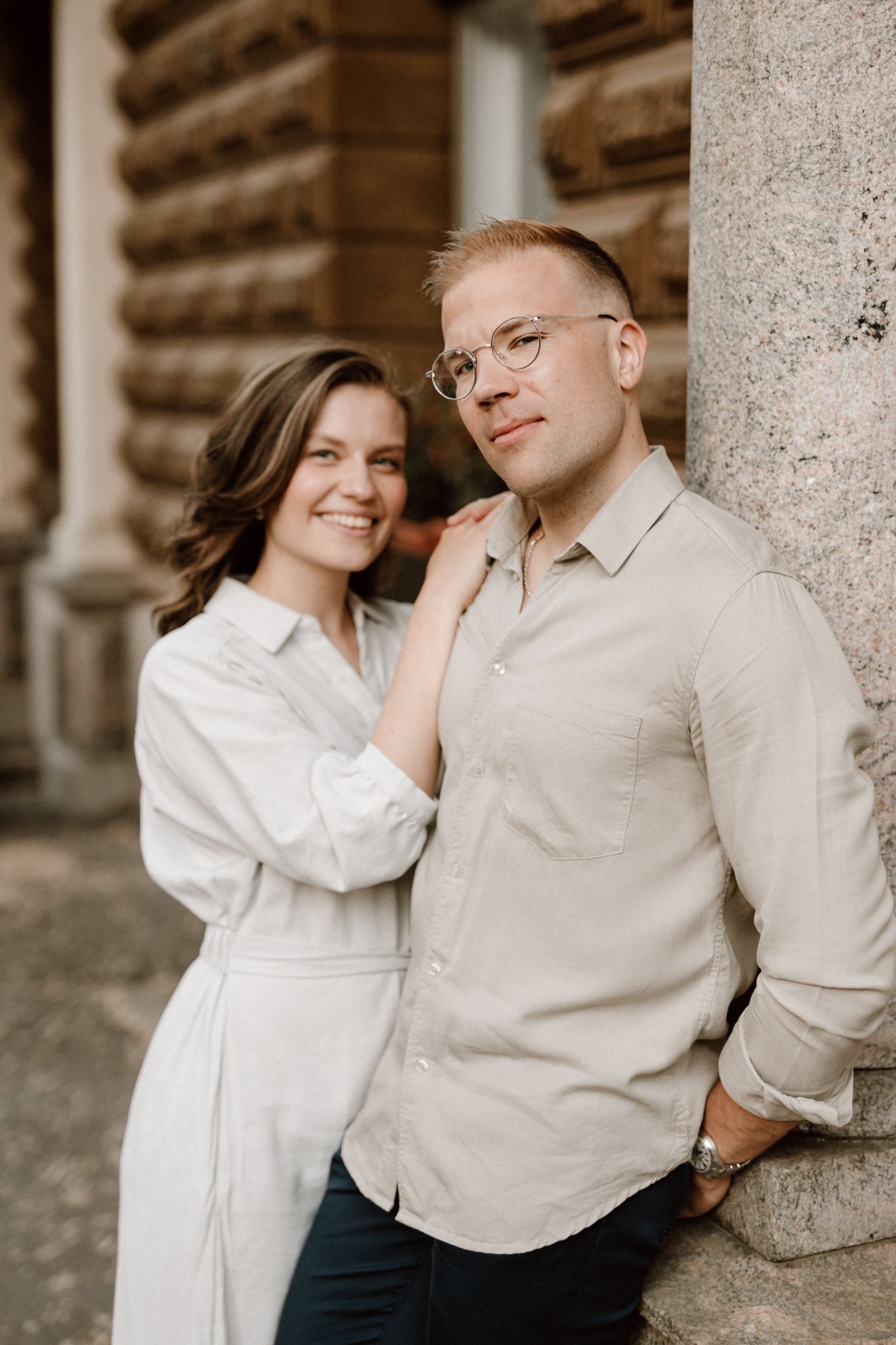 Smiling woman in white dress stands beside a man in beige shirt leaning against a stone column.