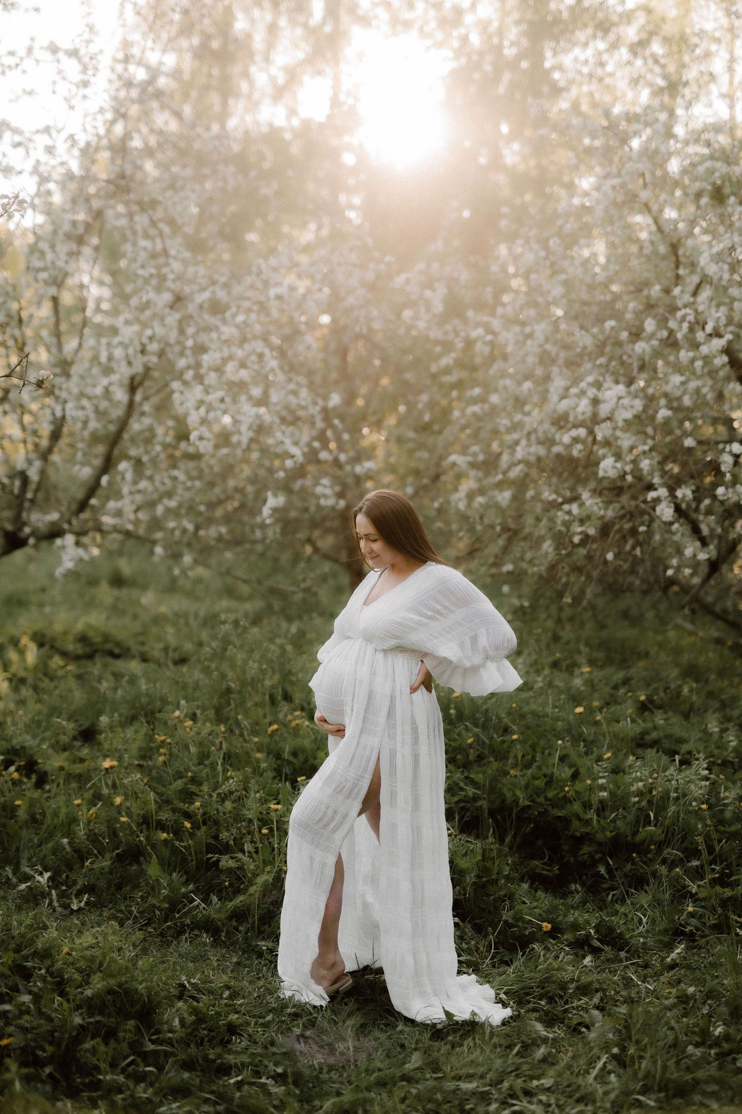 Pregnant woman in elegant white lace dress cradling baby bump among flowering trees in warm sunset glow.