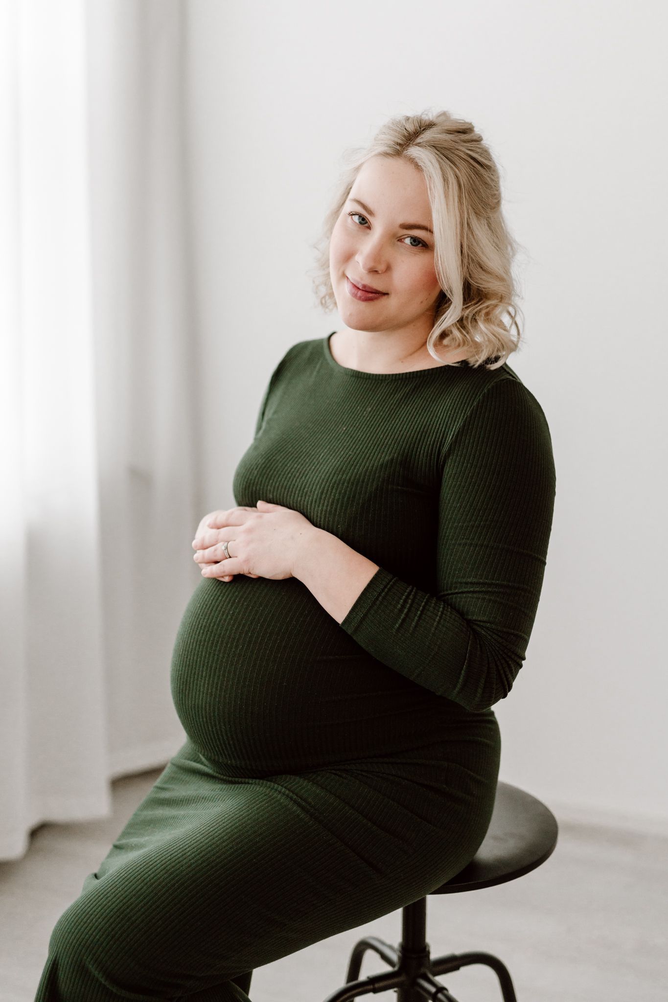 Smiling pregnant blonde woman in dark green ribbed dress sits on stool cradling her baby bump.