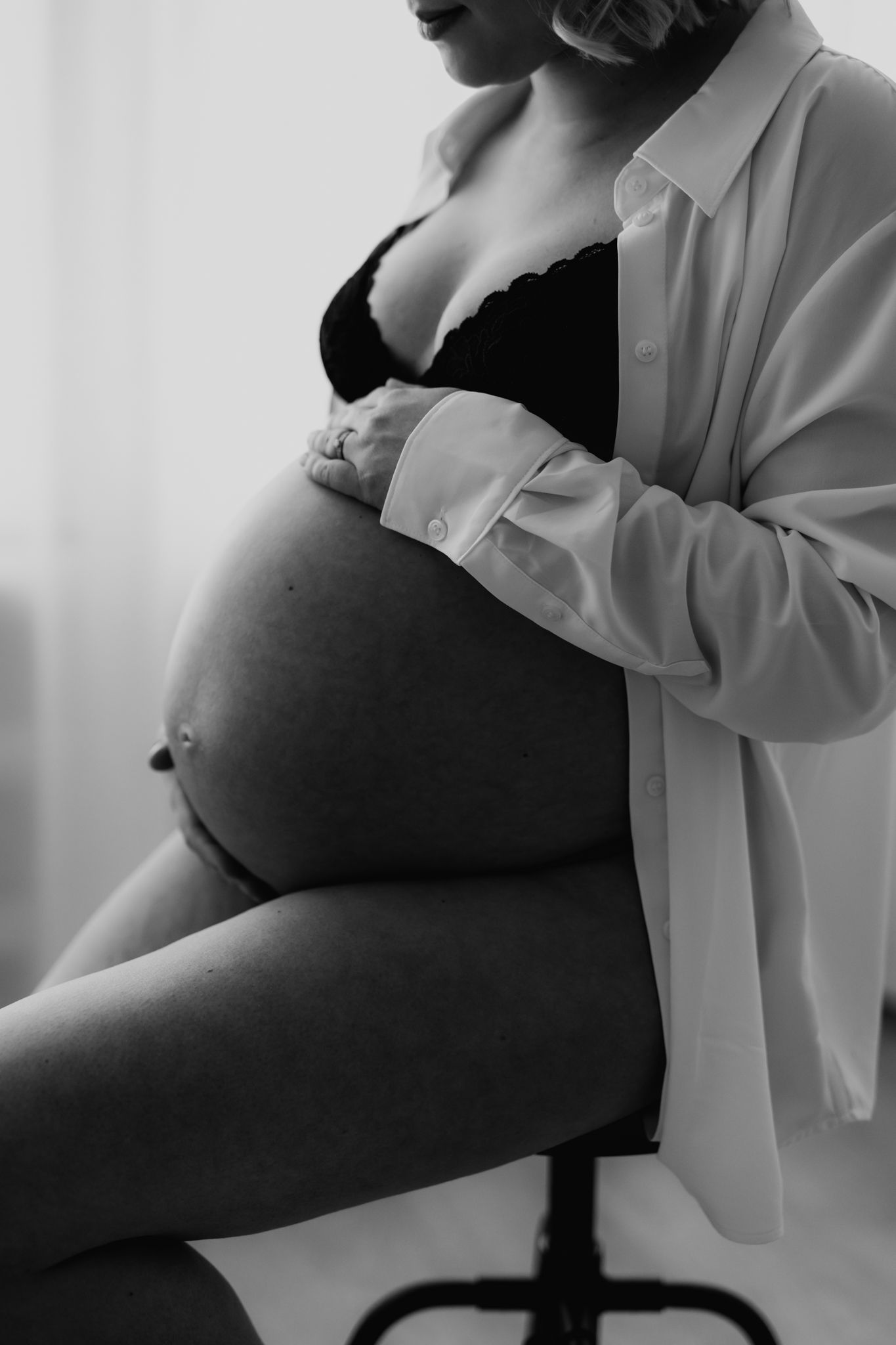 Black and white close-up of pregnant woman in black bra and white shirt sitting and holding bump.