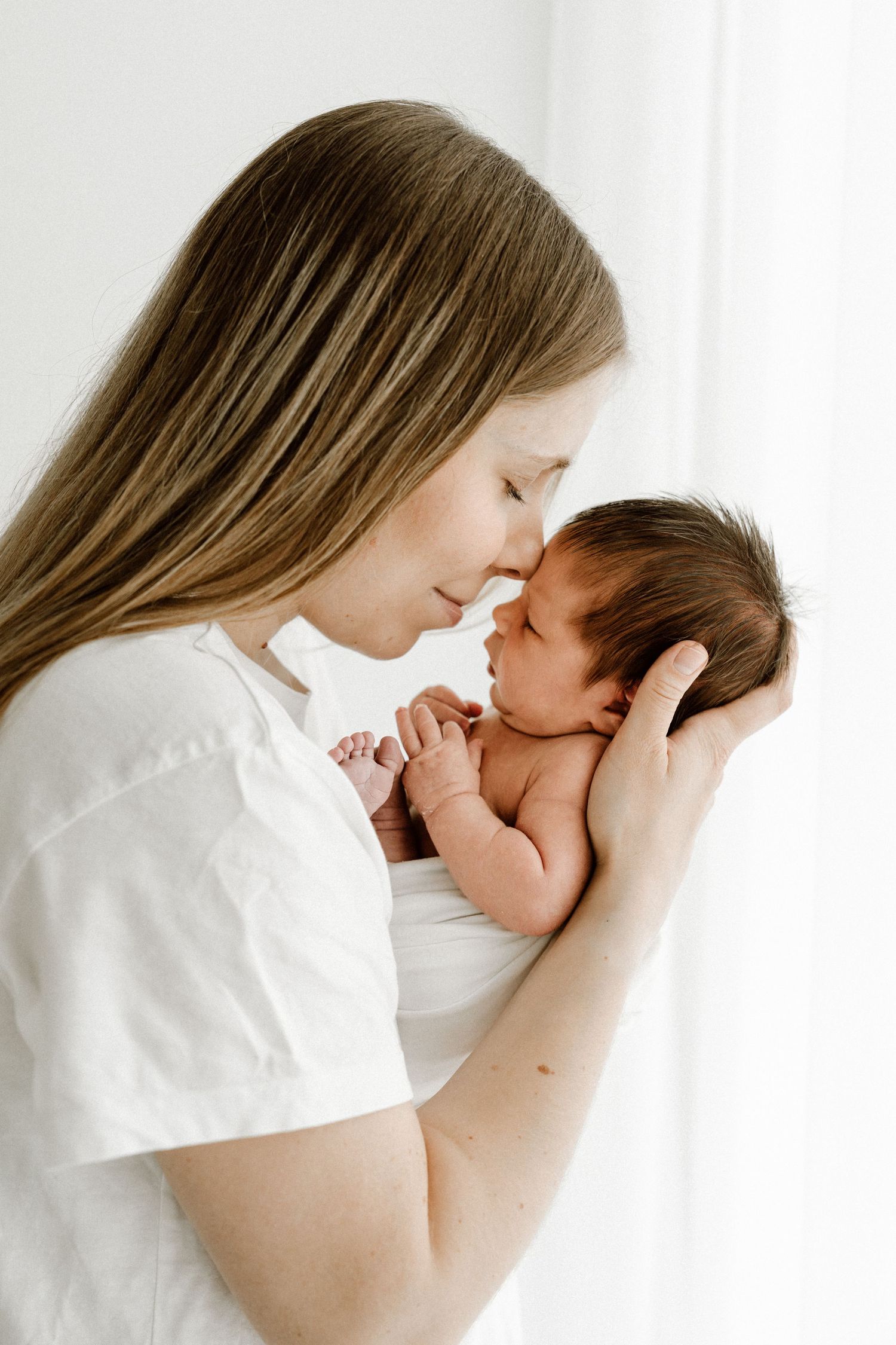 Mother with long hair gently touching foreheads with her swaddled newborn baby.