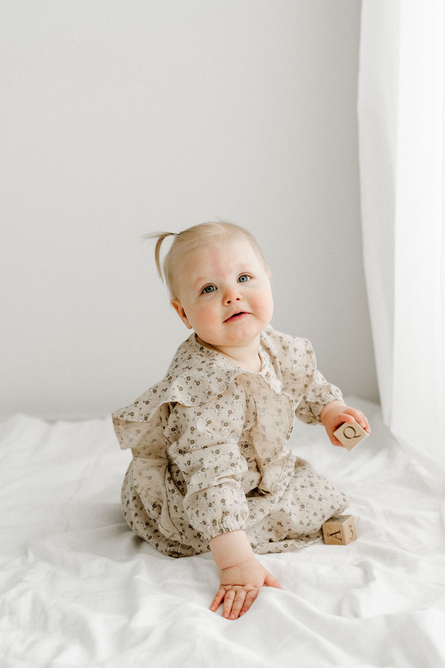 An adorable baby girl in a floral romper sits on a white blanket holding a wooden block, looking up curiously.