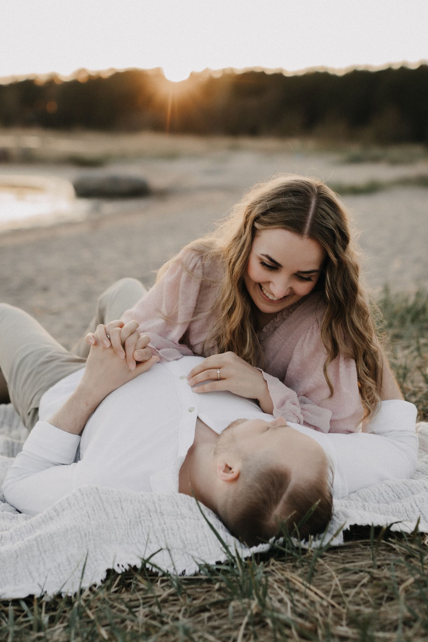 Couple relaxing on blanket at sunset, woman in pink dress smiling while man lies on her lap.
