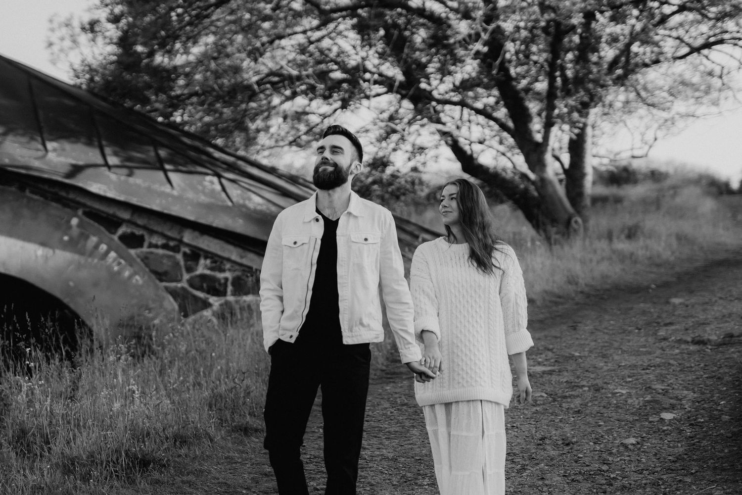 Black and white photo of a couple holding hands walking near rustic stone wall outdoors.