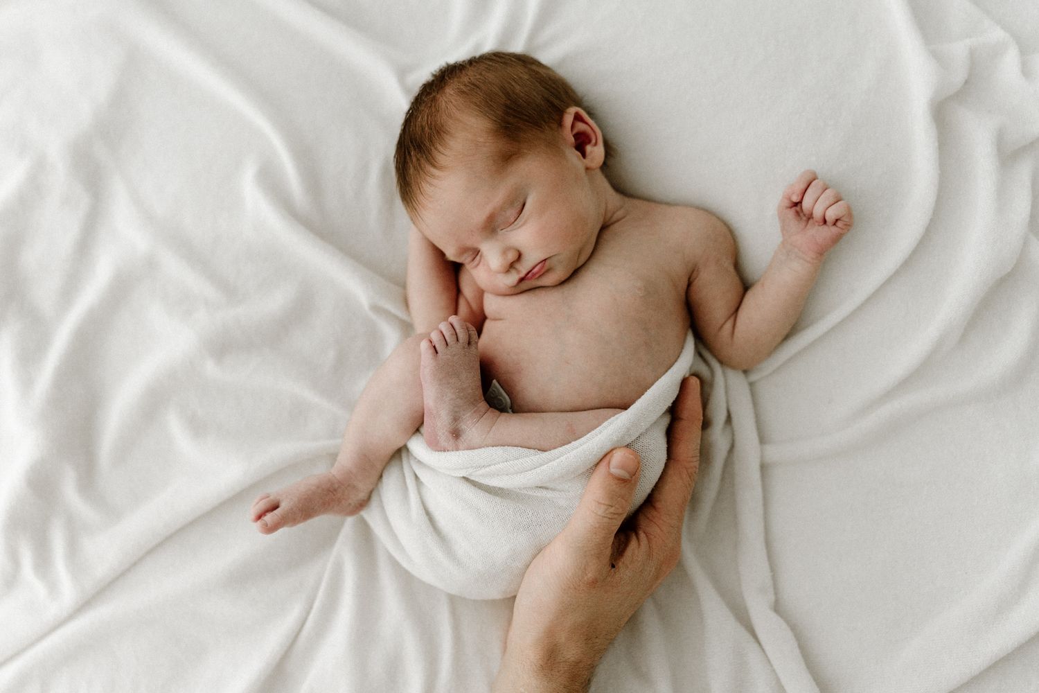 Adult hands gently swaddling a peacefully sleeping newborn baby in white wrap.
