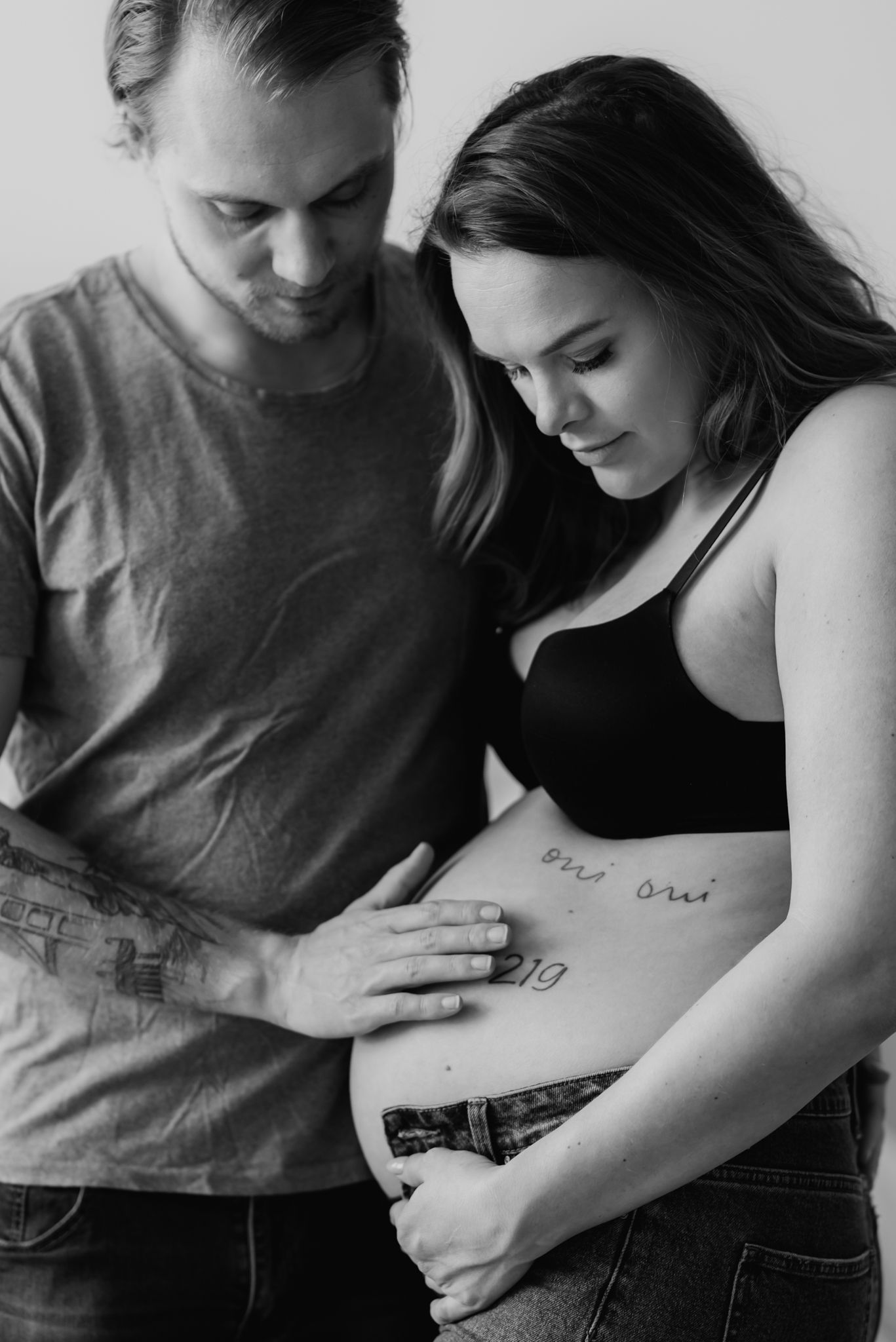 Black and white maternity photo of couple tenderly looking at pregnant belly with tattoos.