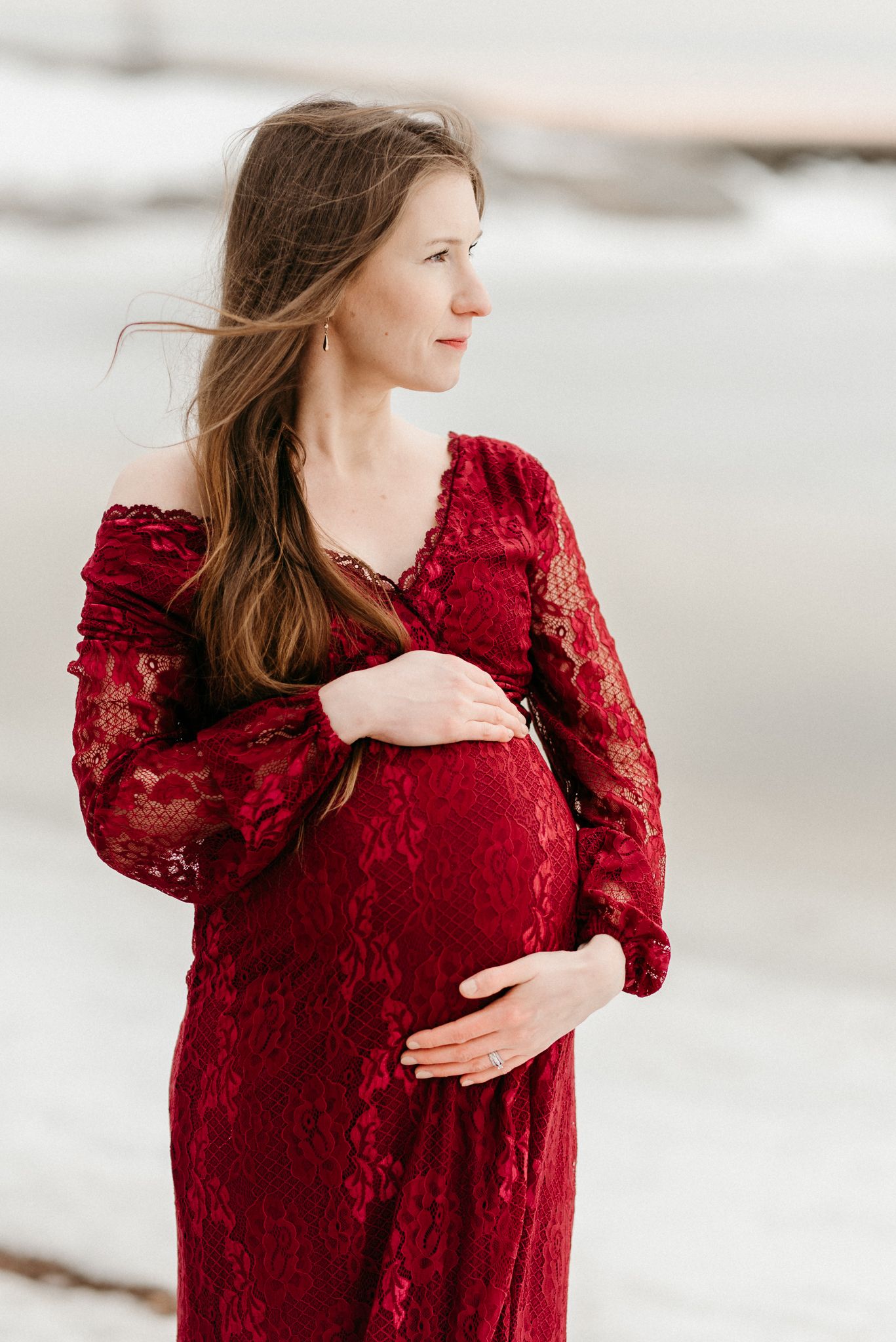 Pregnant woman in elegant red lace off-shoulder gown cradling baby bump outdoors near snowy winter shoreline.