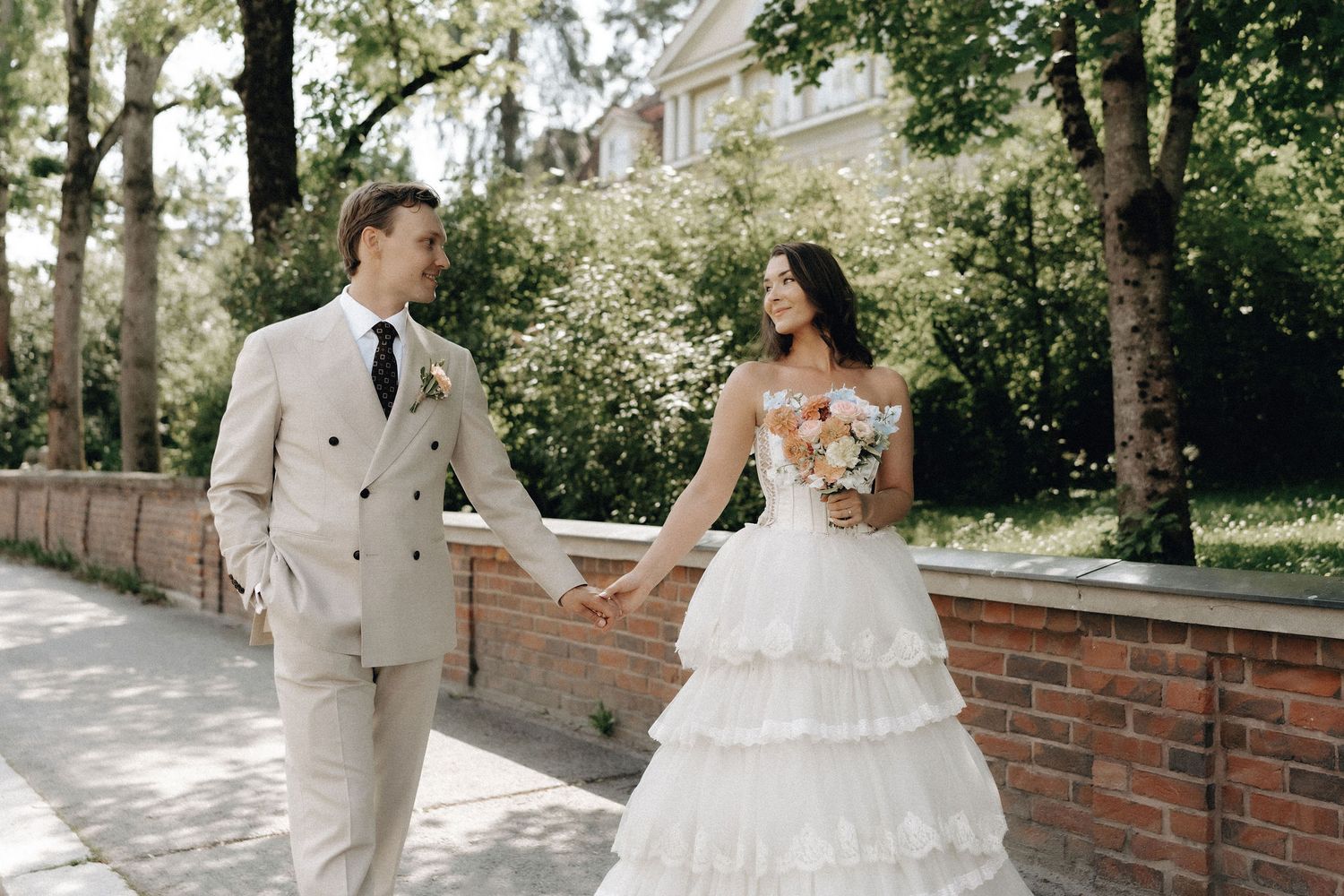 Bride in tiered white gown and groom in beige suit hold hands walking along a tree-lined path.