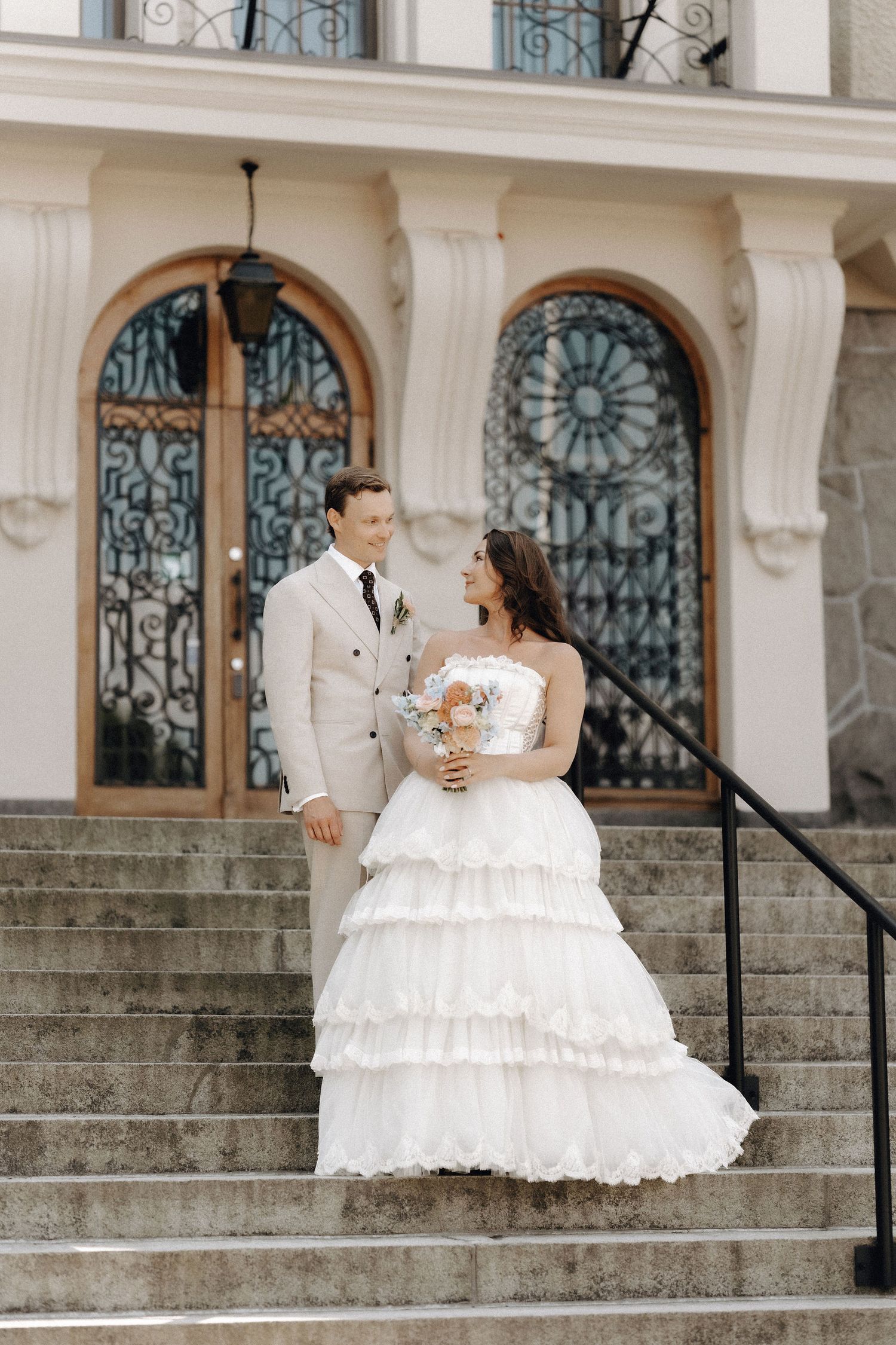Bride and groom pose on stone steps before ornate building with arched stained glass doors.