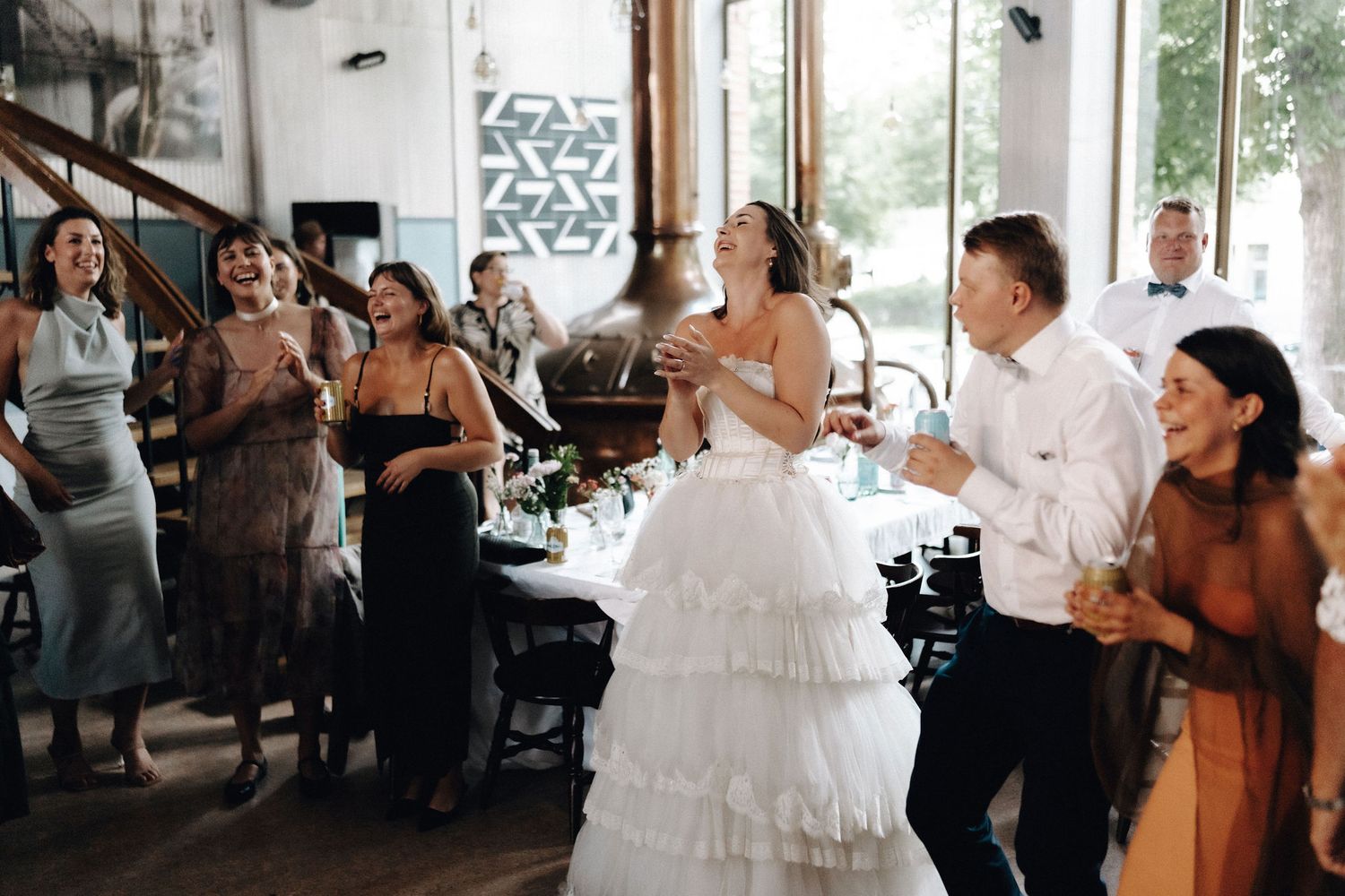 Bride in ruffled white gown laughing with groom and guests at wedding reception.