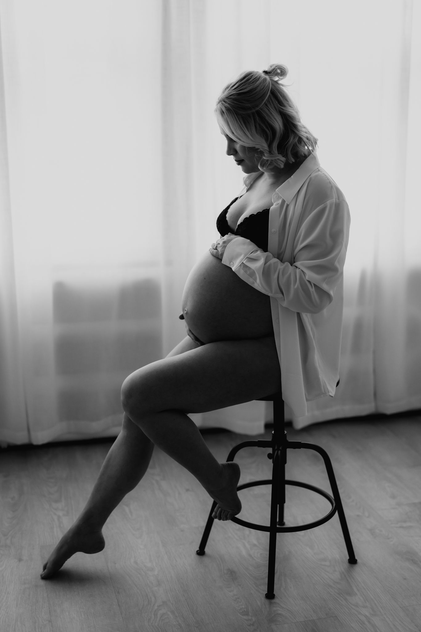 Black and white silhouette of pregnant woman in lingerie and open white shirt sitting on stool.