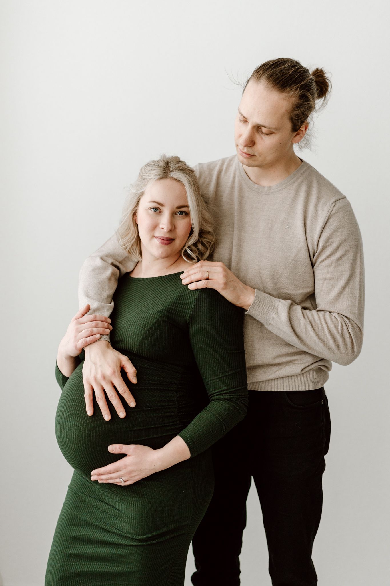 Pregnant woman in green dress embraced from behind by partner against light gray background.