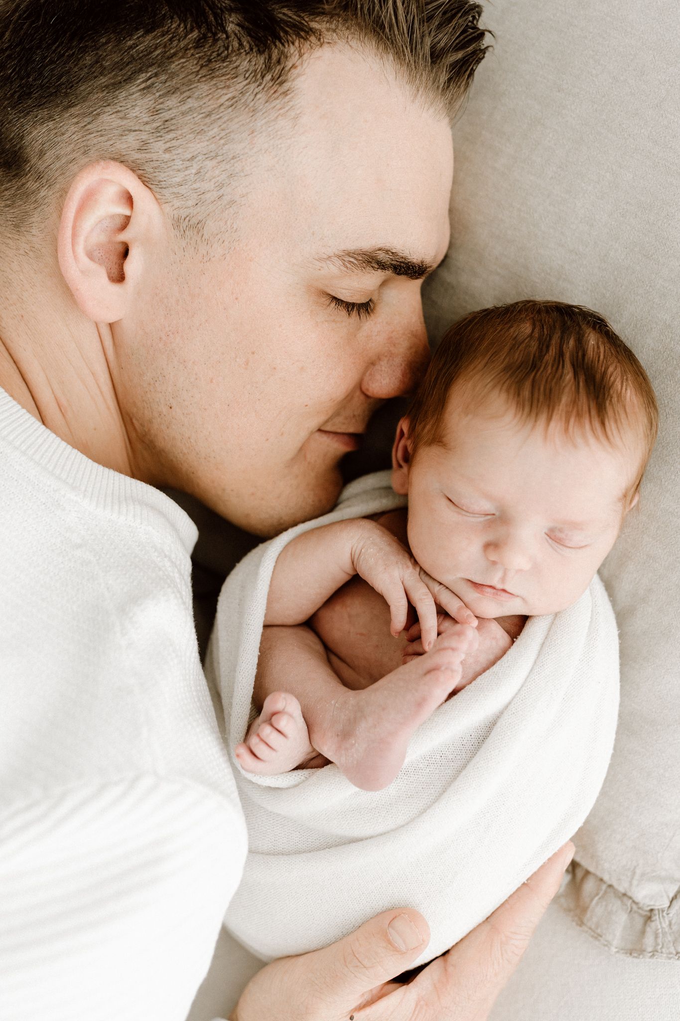 Father tenderly nuzzles sleeping newborn baby wrapped in white swaddle blanket.