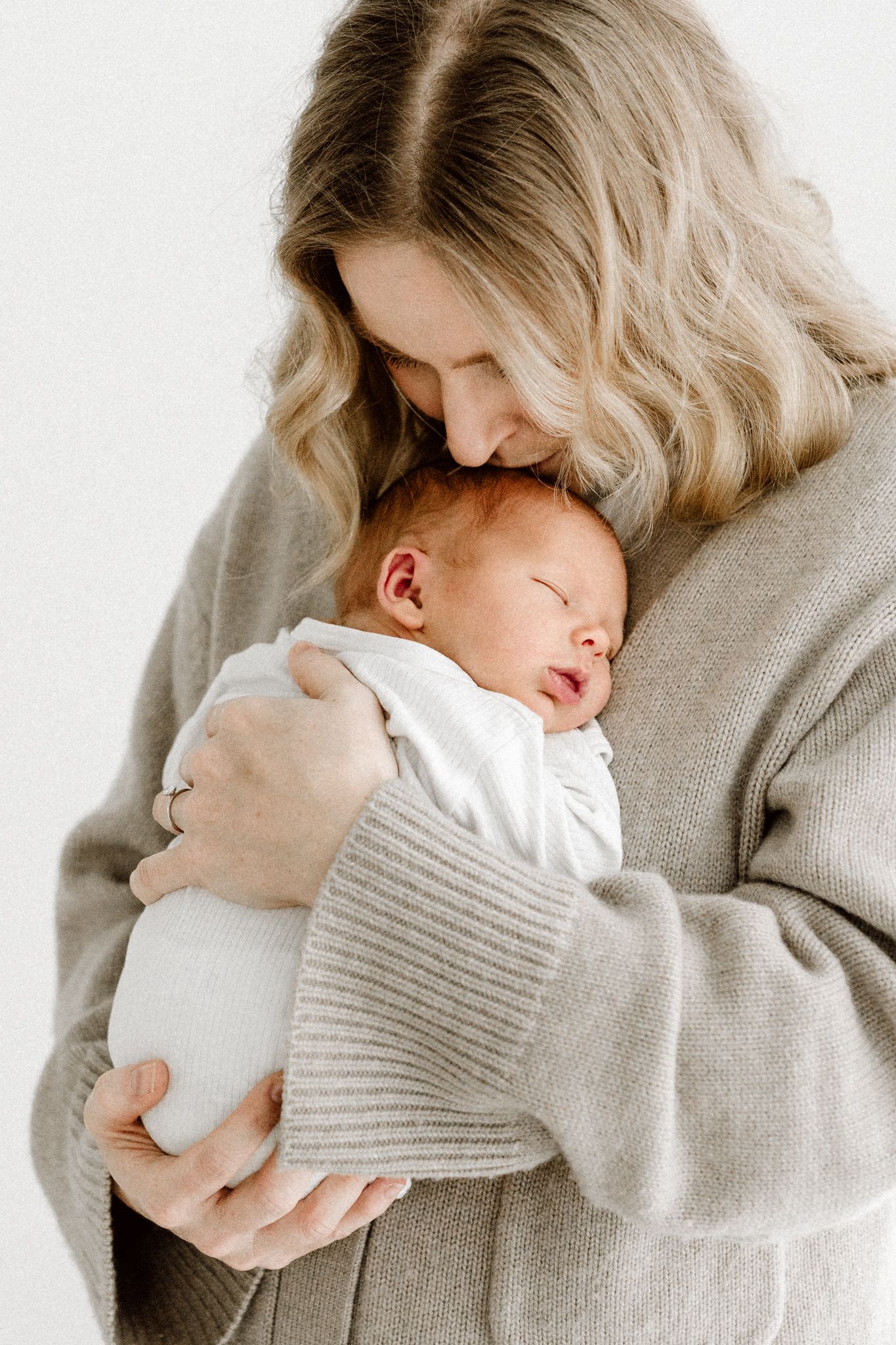 Blonde mother in beige cardigan tenderly nuzzling her sleeping newborn baby close to her chest.