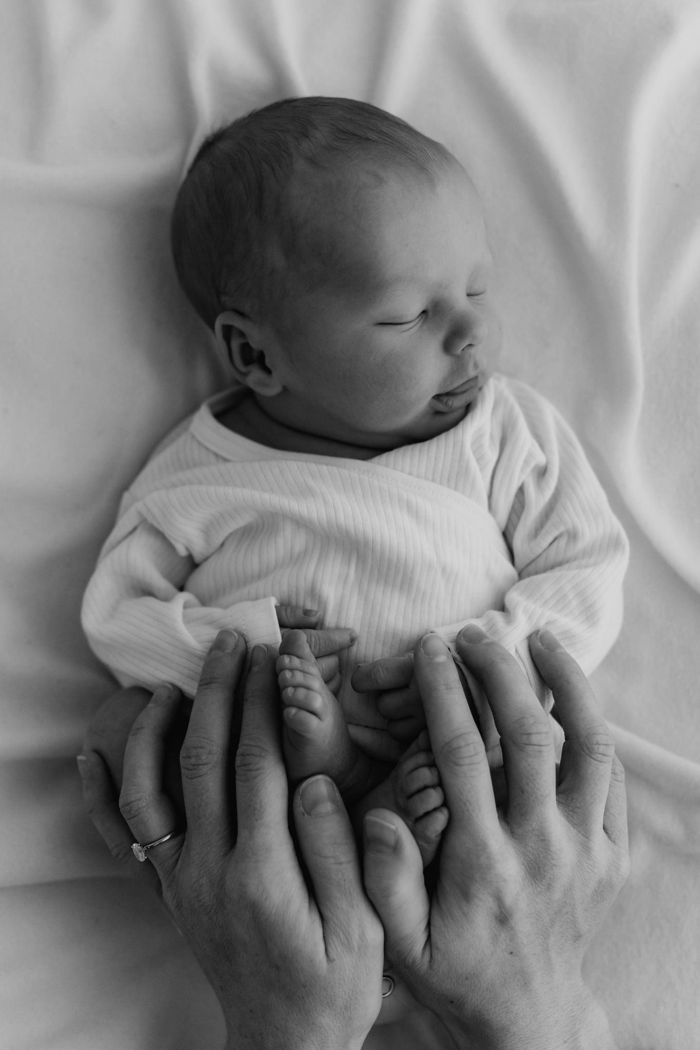 Black and white overhead photo of a peacefully sleeping newborn with parents hands gently holding tiny feet.