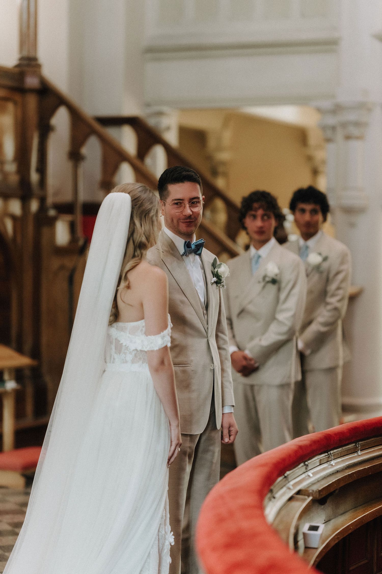 Groom in tan suit smiling at bride in white wedding dress and veil at church altar with groomsmen standing behind.