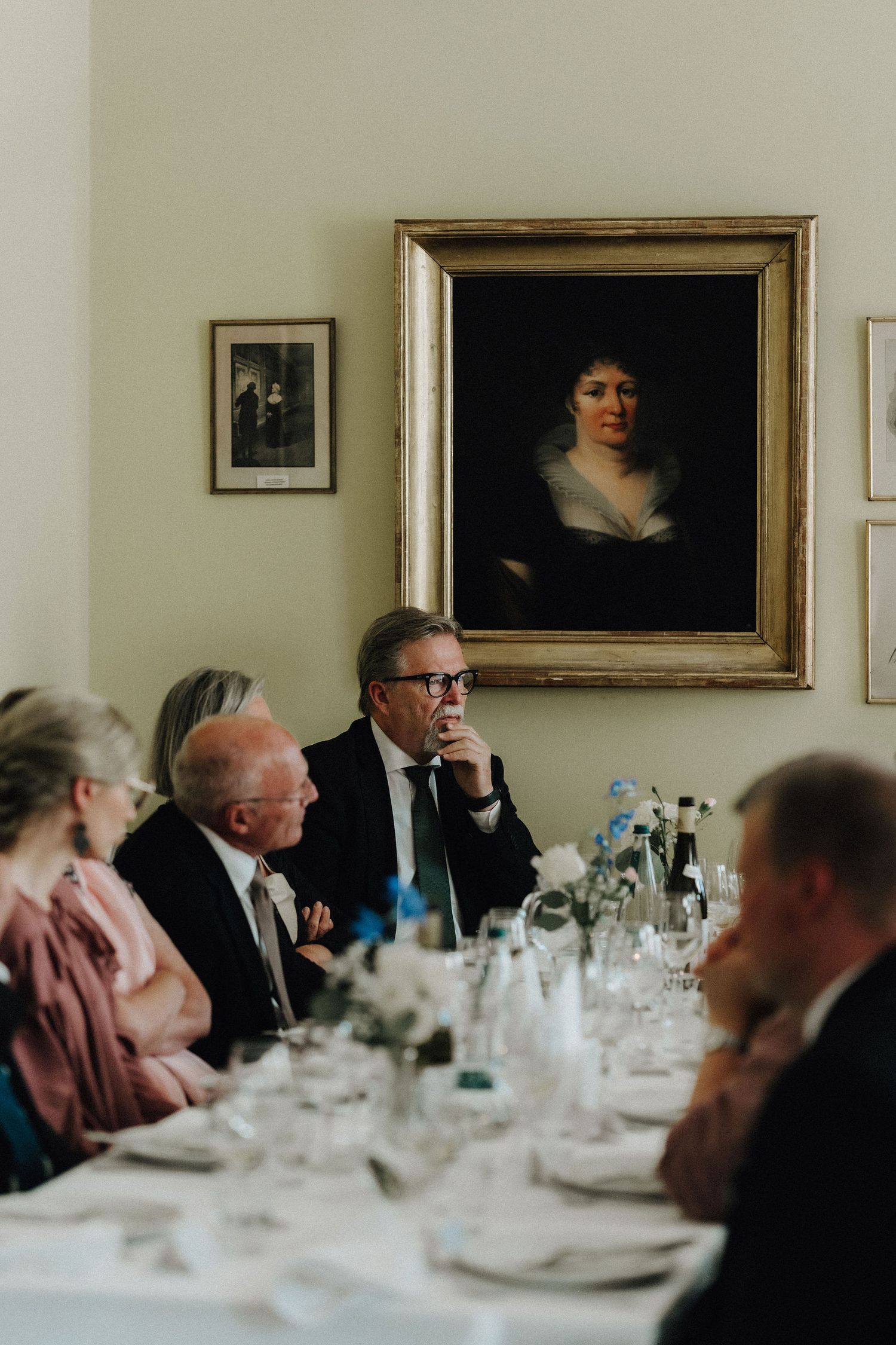 Wedding guests seated at a formal dinner table beneath a classic portrait painting on a cream wall.
