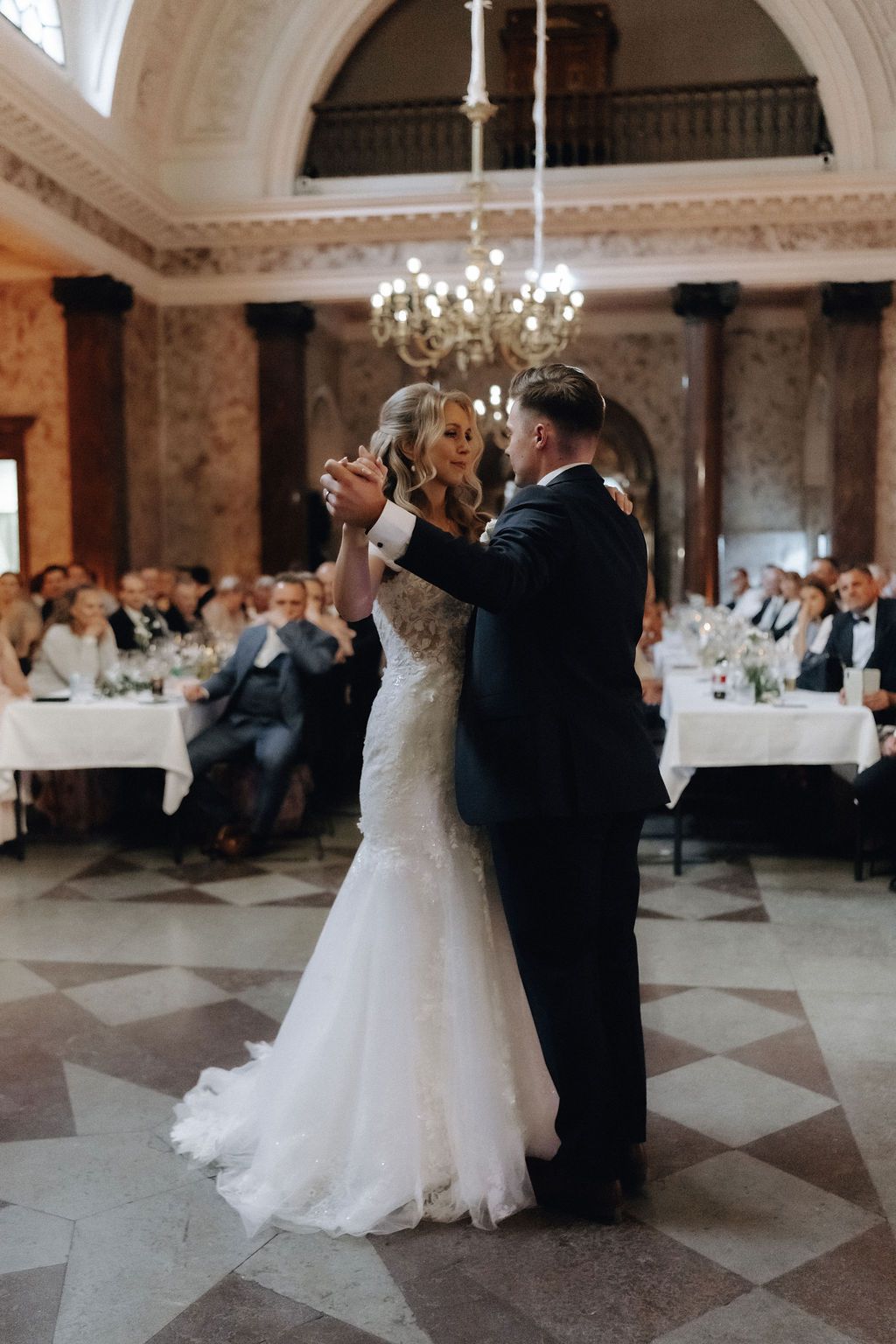 Bride and groom share romantic first dance under chandelier in grand arched marble ballroom.