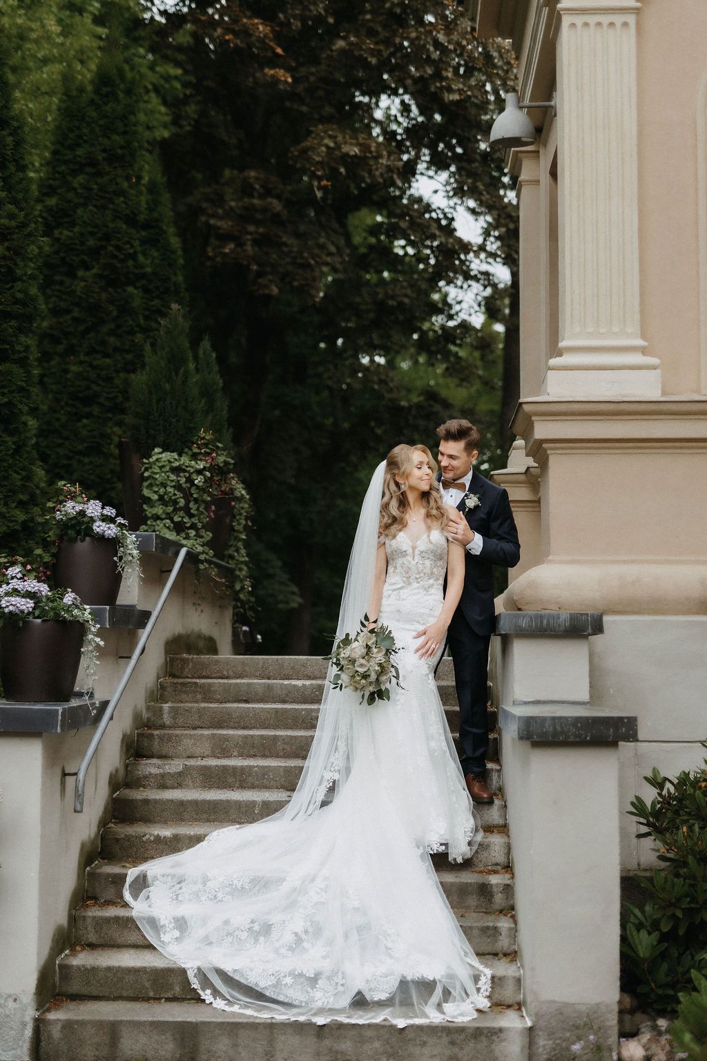 Bride and groom pose on elegant stone steps outside a classical building surrounded by lush greenery.