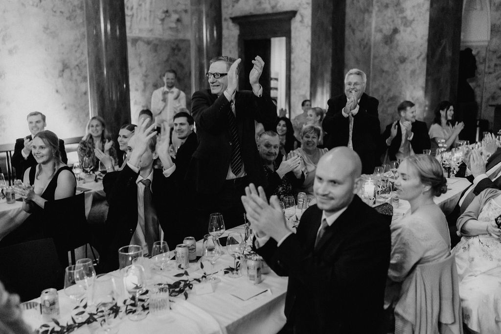 Wedding guests clap and celebrate during reception dinner in elegant marble ballroom.
