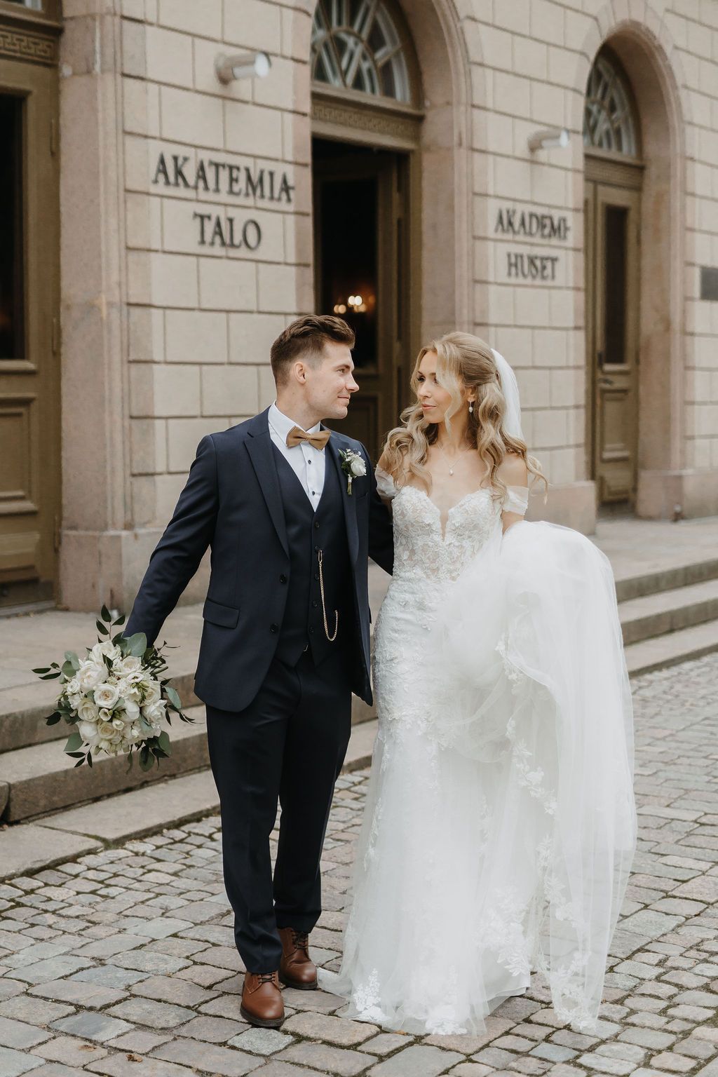 Bride and groom walk together smiling outside Akatemia Talo, she in lace gown, he in navy suit.