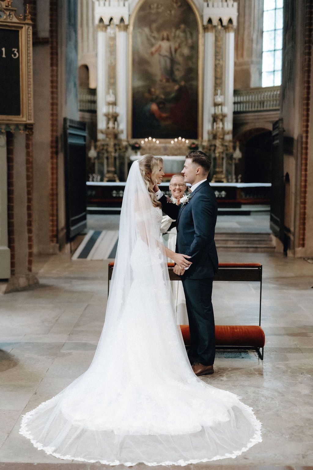 Bride and groom hold hands before officiant at ornate church altar during wedding ceremony.