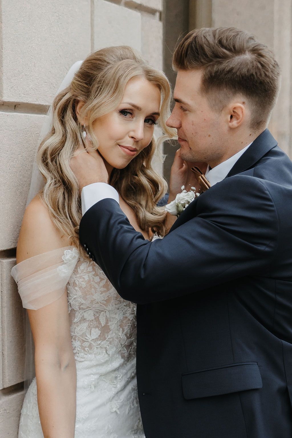 Groom tenderly touching bride's face as she smiles against a stone wall on their wedding day.